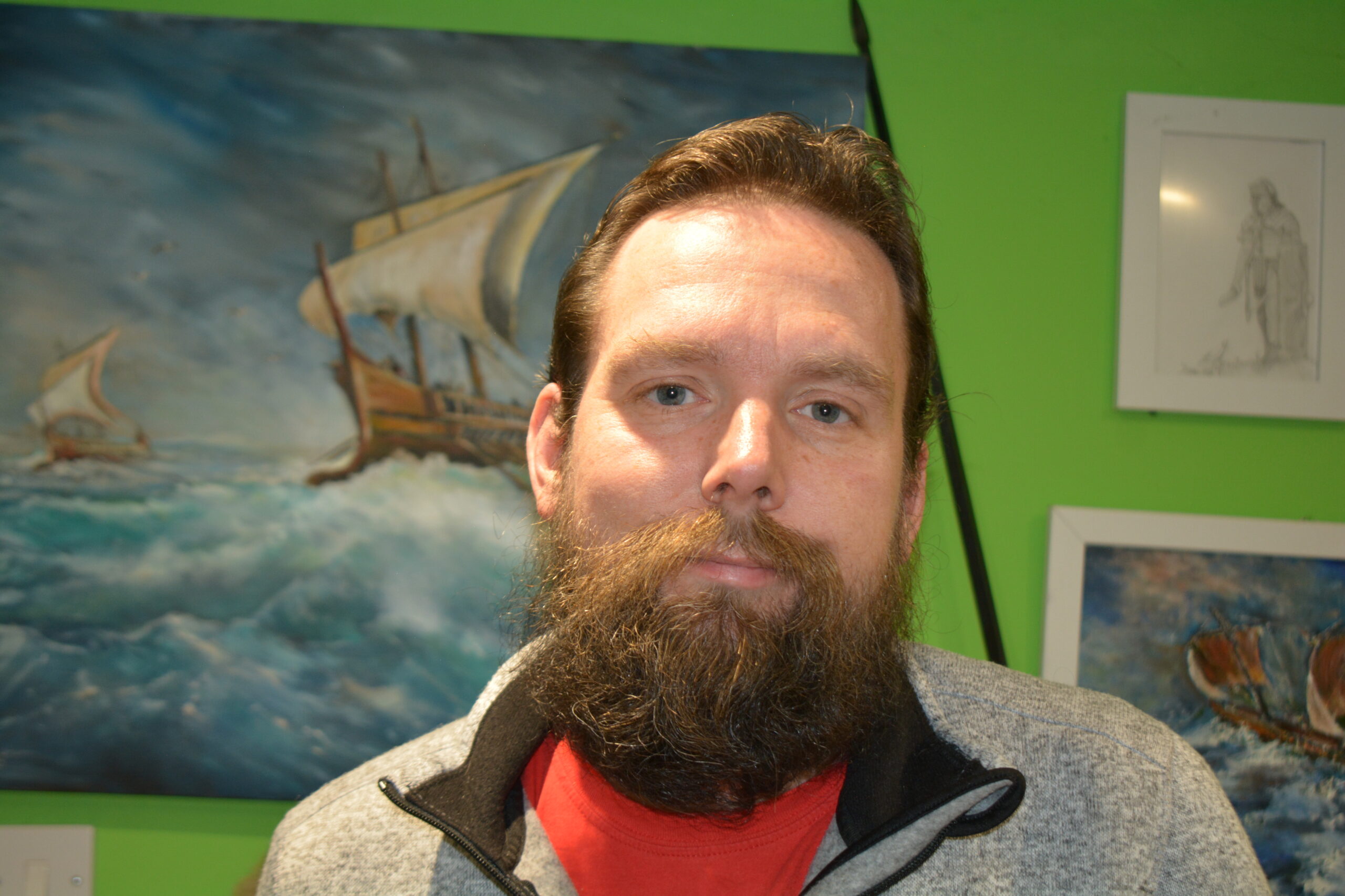 A man with a beard is standing in front of a colorful wall adorned with nautical artwork. He wears a gray zip-up jacket over a red shirt, looking directly at the camera with a neutral expression.