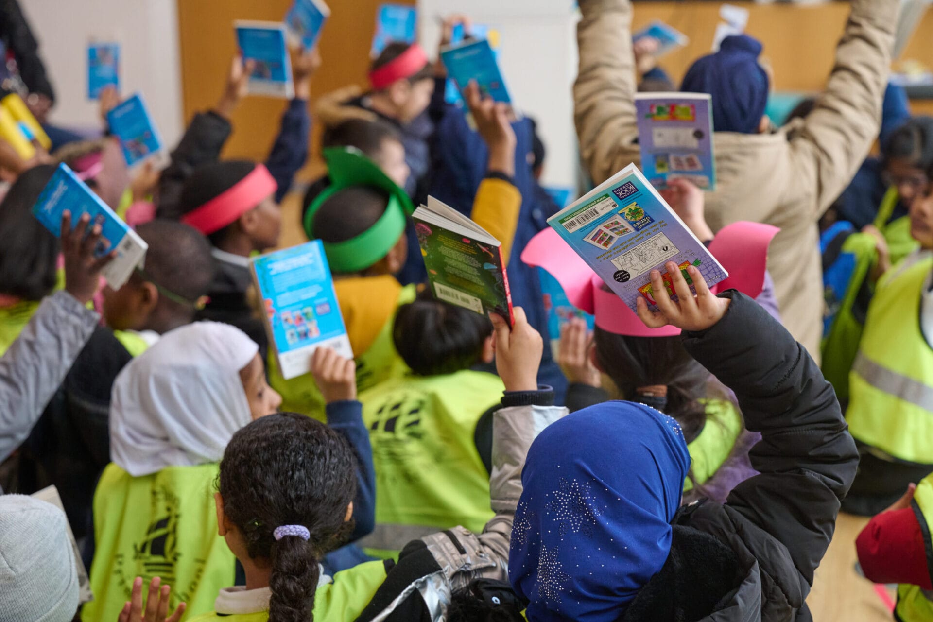 A group of children in colorful headbands enthusiastically raise books in a classroom setting, showcasing participation and engagement in a reading activity or discussion.