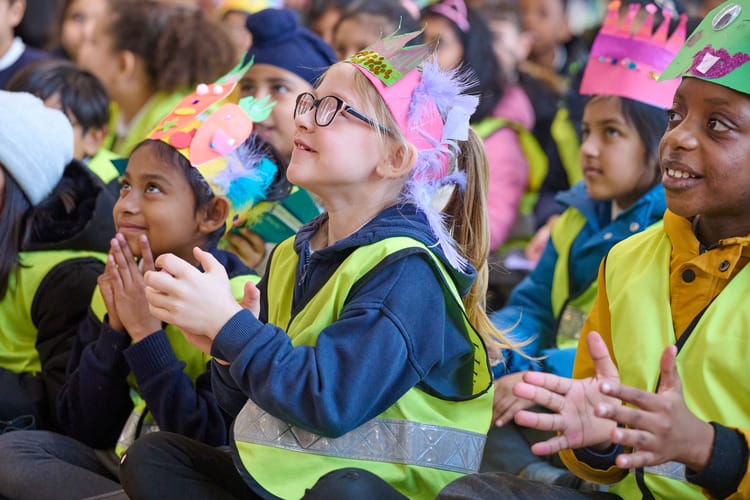 A group of diverse children, wearing bright safety vests and colorful paper crowns, sit together, smiling and clapping enthusiastically. They are engaged in an activity, showcasing joy and excitement in a lively, participatory setting.