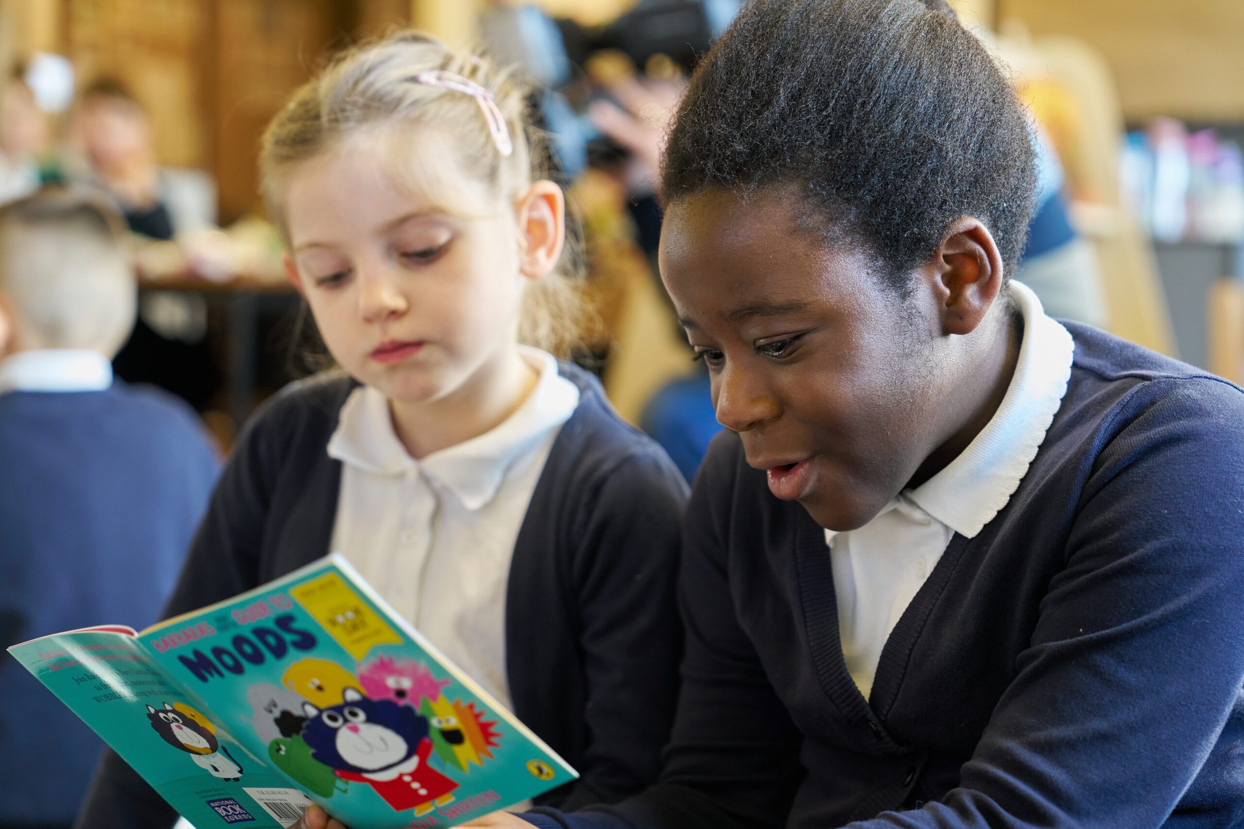 Two children are seated together, one reading a colorful book titled "Moods." The girl on the left looks on with interest, while the boy on the right expresses excitement as he engages with the story. The setting suggests a classroom environment.