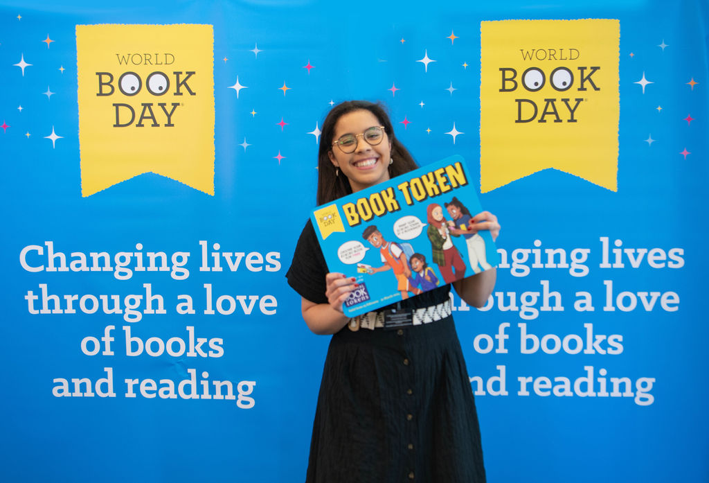 A smiling individual holds a "Book Token" in front of a vibrant background that highlights World Book Day and emphasizes the theme of changing lives through a love of books and reading.