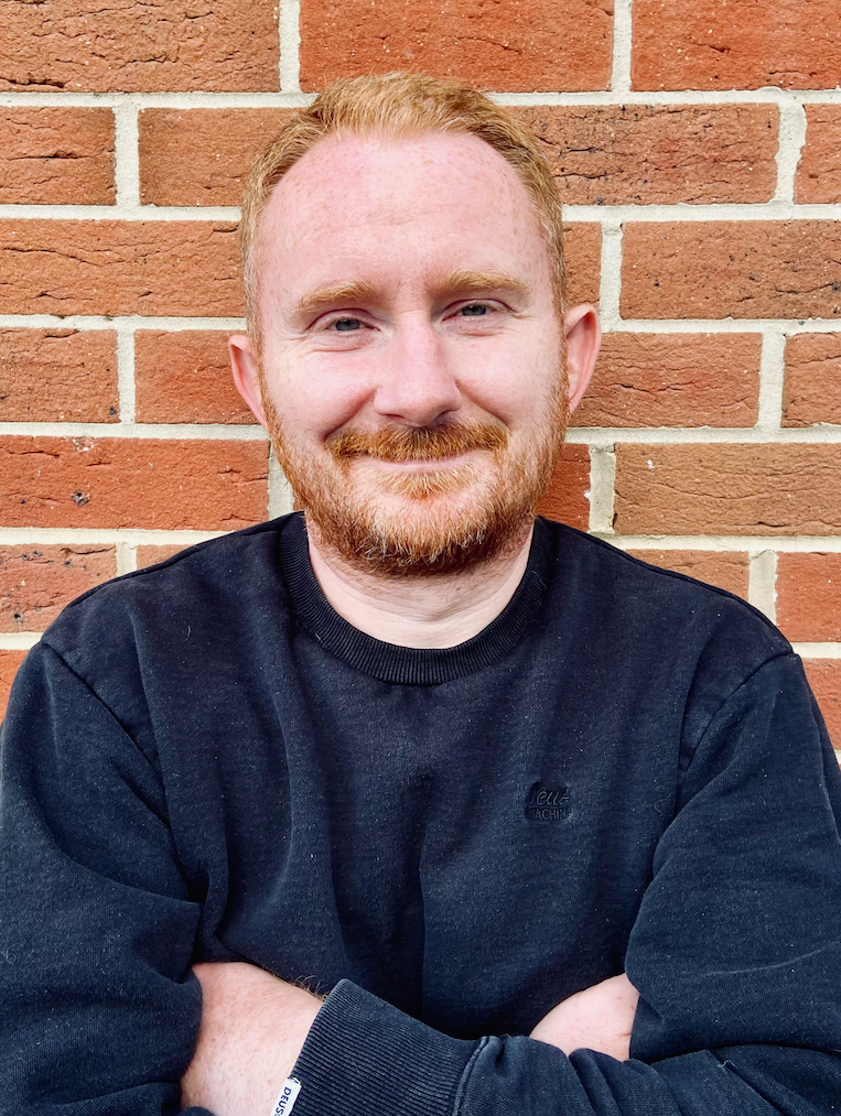 A smiling man with red hair and a beard poses with crossed arms against a red brick wall, wearing a black sweatshirt.