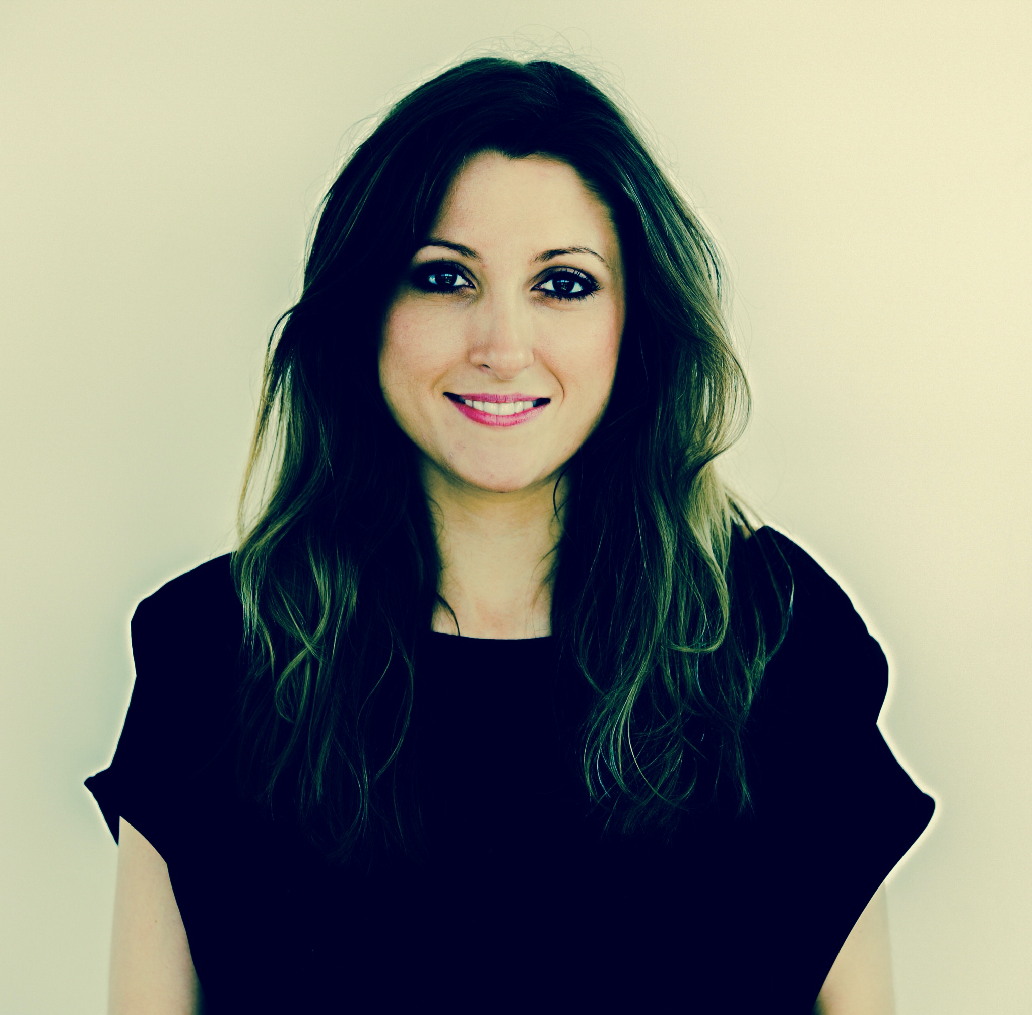 A woman with long, wavy hair and a slight smile stands against a light background, wearing a black top. Her expression is friendly and approachable, conveying warmth and confidence.