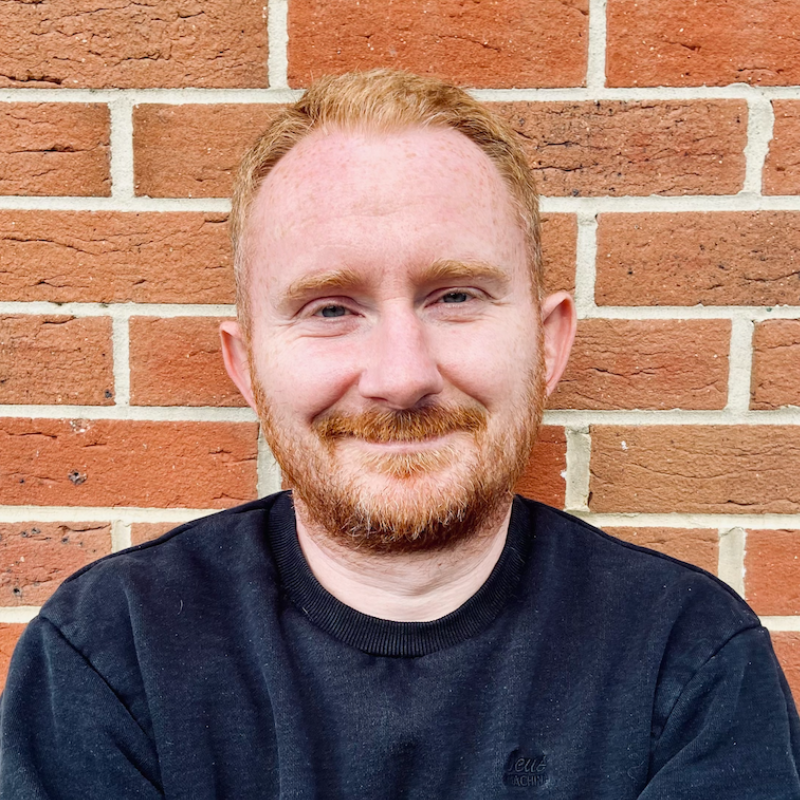A person with short red hair and a beard smiles while leaning against a brick wall. They are wearing a black sweatshirt. The background features a textured pattern of red and beige bricks.