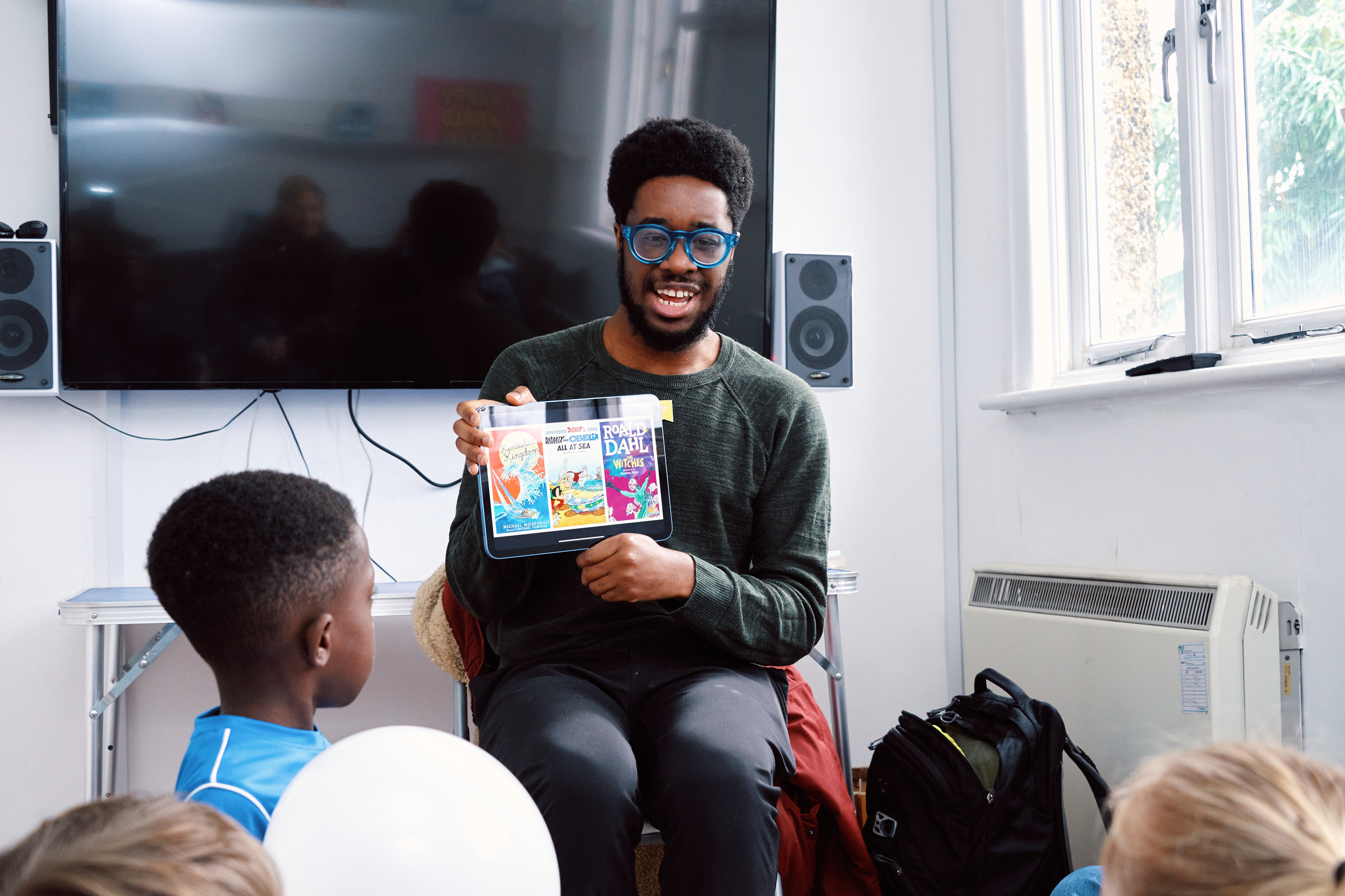 A smiling man with glasses holds a colorful box of books, engaging a group of children seated around him. The setting features a bright room with a large display screen in the background, creating an inviting atmosphere for storytelling.
