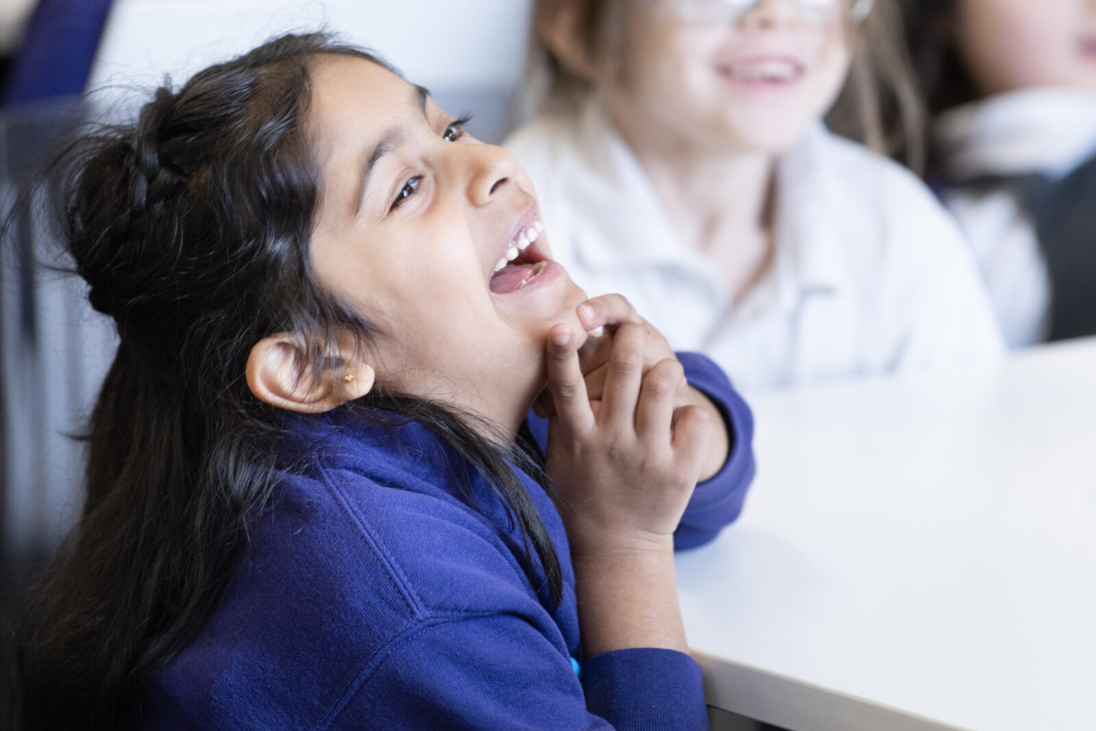 A young girl with black hair laughs joyfully while sitting at a table, with her hands clasped under her chin. Other children are visible in the background, also engaged and smiling.