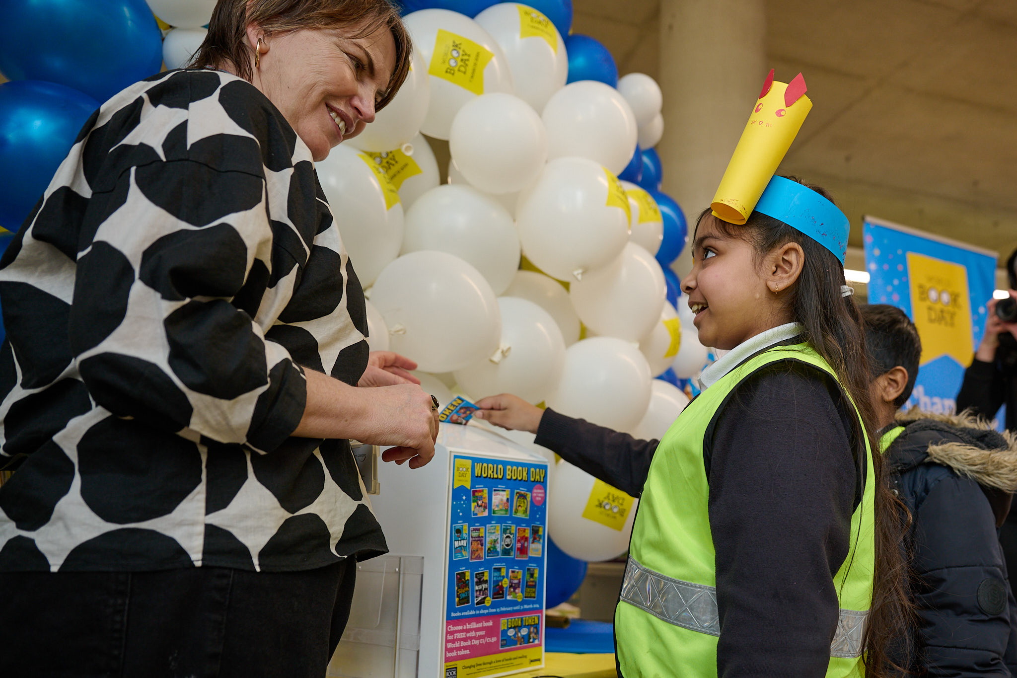 A woman interacts with a smiling child wearing a colorful crown and a bright vest at an event. Balloons decorate the background, and a display for World Book Day is visible nearby, emphasizing a celebration of reading and literacy.