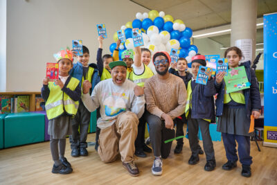 A group of cheerful children in bright vests gather with two adults, smiling and holding up books. They are celebrating a reading event, surrounded by colorful balloons and a vibrant backdrop.
