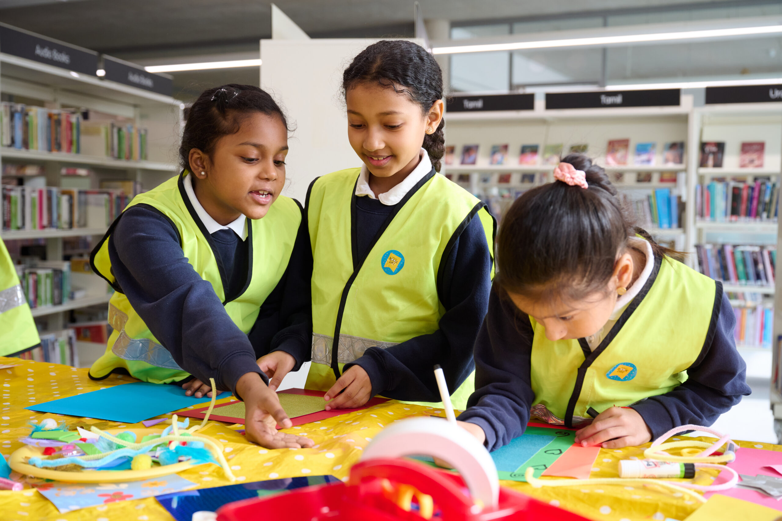 Three children wearing bright yellow vests are engaged in a craft activity at a table filled with colorful materials in a library. They are focused on creating art while surrounded by shelves of books.