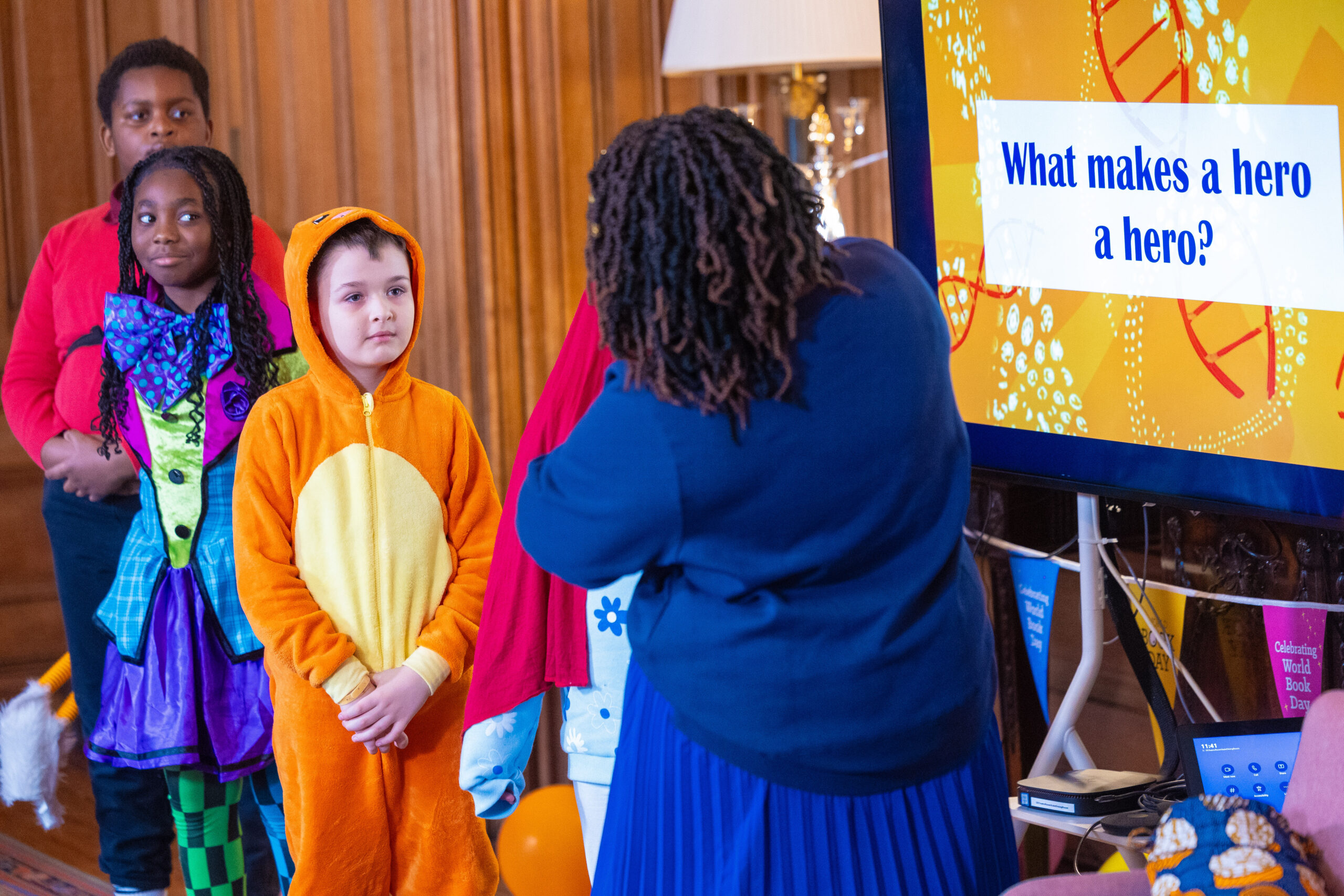 A group of children in colorful costumes listens to an adult speaker, who is posing the question, "What makes a hero a hero?" The setting appears festive, likely during a themed event or celebration.