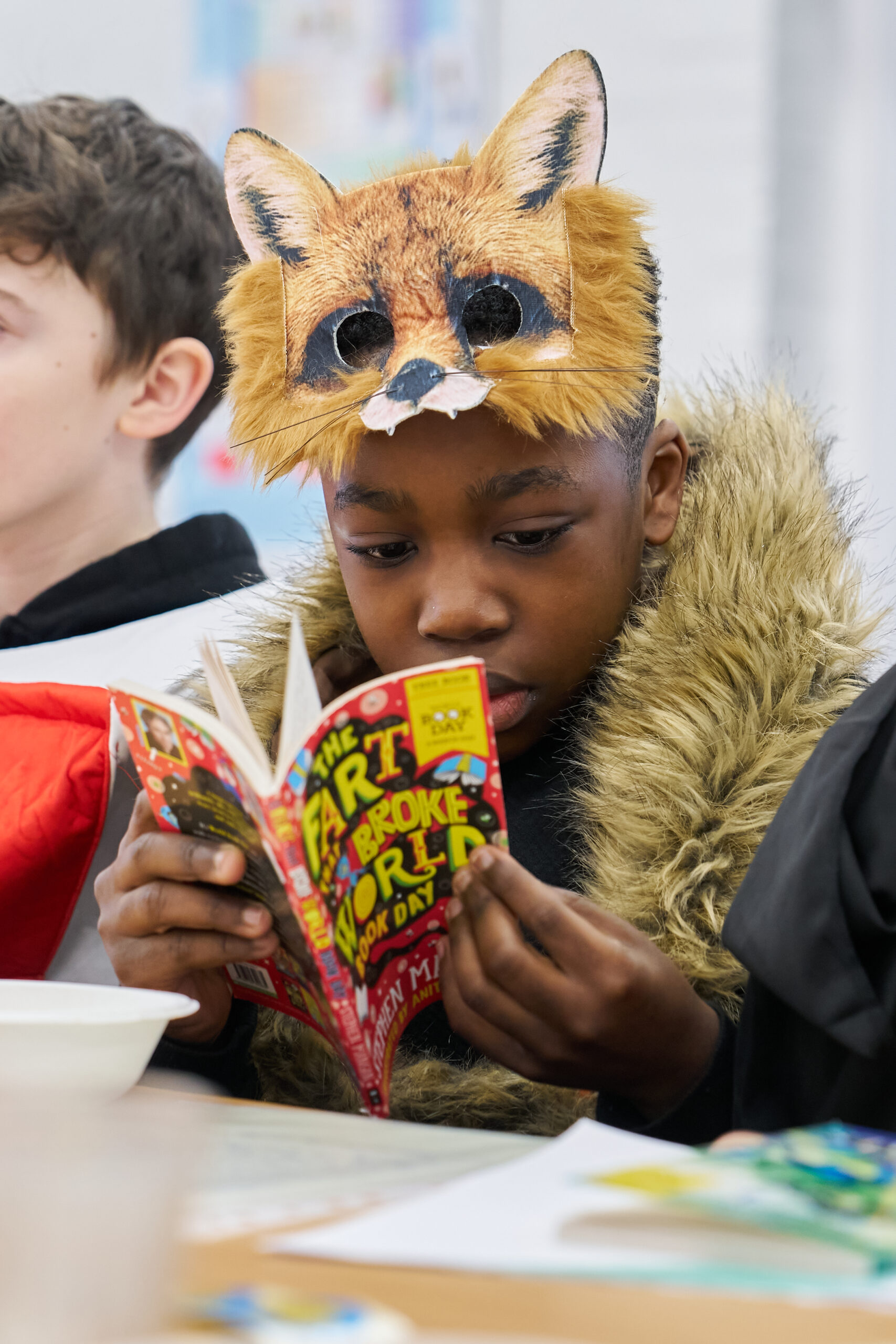 A child wearing a fox hat reads a colorful book titled "The Fart That Broke the World." They are seated at a table with other children, focused on their reading while dressed in a furry coat.
