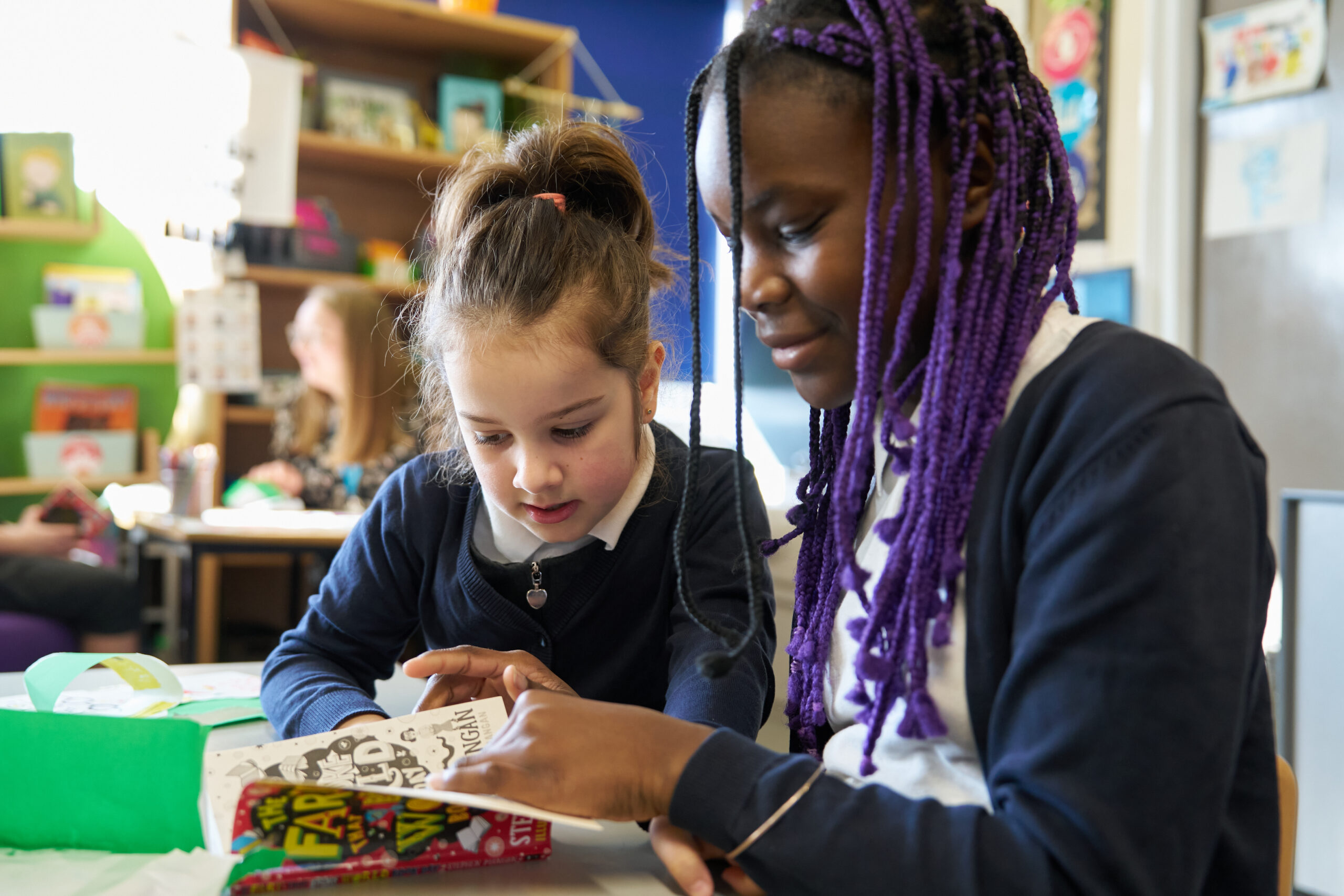 Two young girls are engaging with a book at a table in a classroom. One girl with purple hair is pointing at the book, while the other girl, with brown hair, looks closely at the pages. Bright classroom decor is visible in the background.