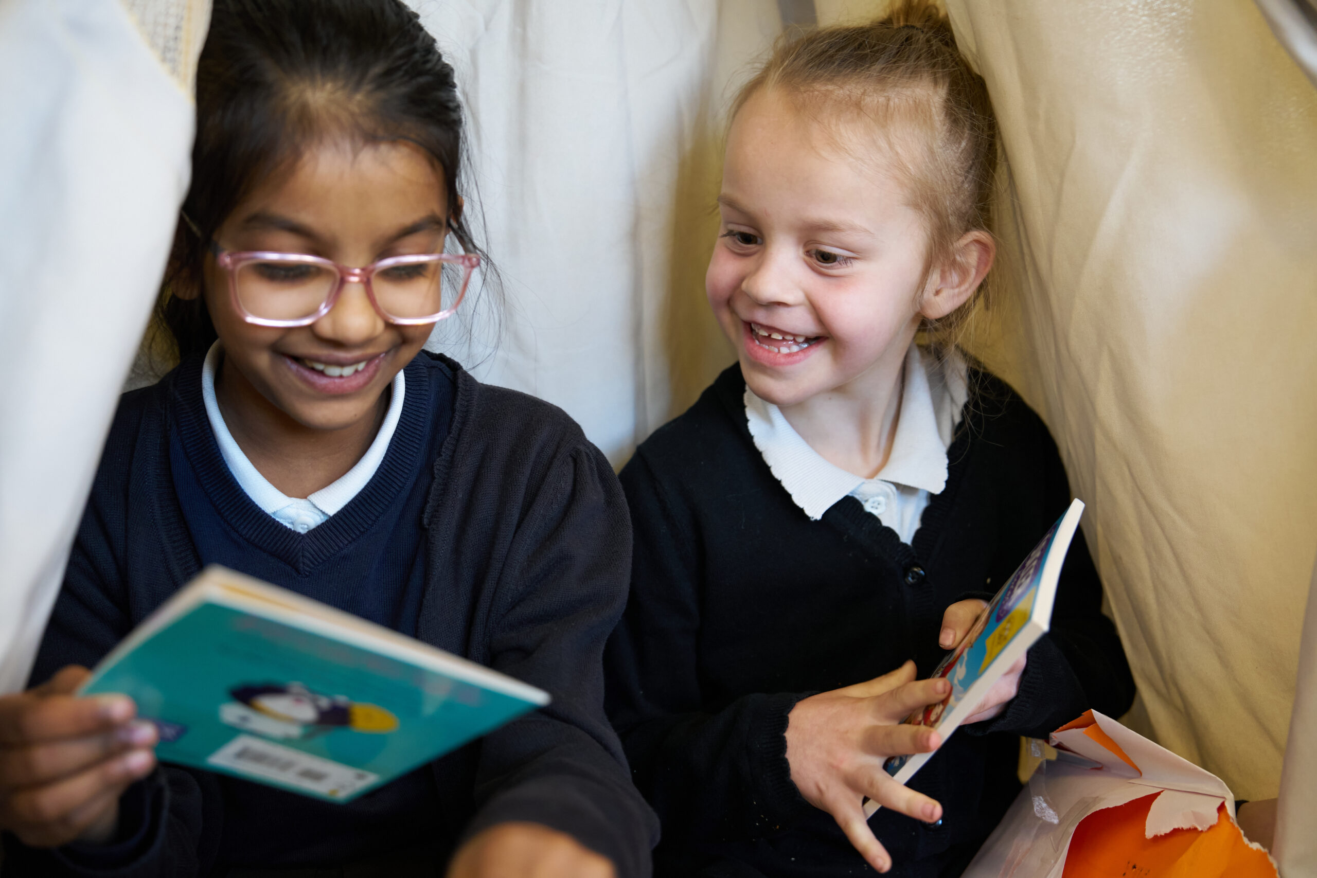 Two young children sit together in a cozy nook, joyfully reading books. One girl wears glasses and smiles broadly, while the other shares a bright grin, both clearly enjoying their reading moment amidst soft fabric surroundings.