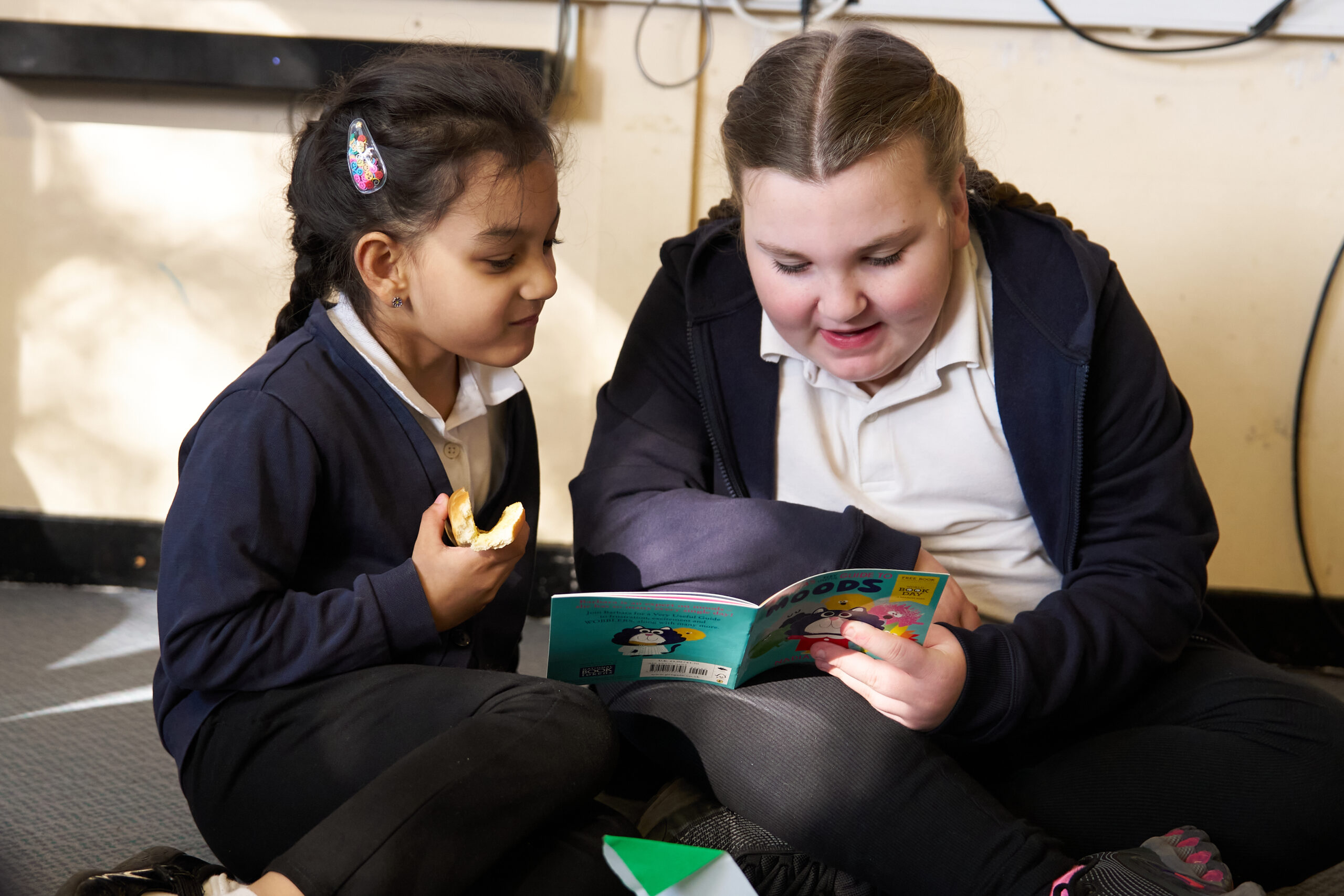 Two girls sit on the floor, closely reading a colorful book together. One girl eats a snack while the other points at the pages, both engaged and enjoying the moment.