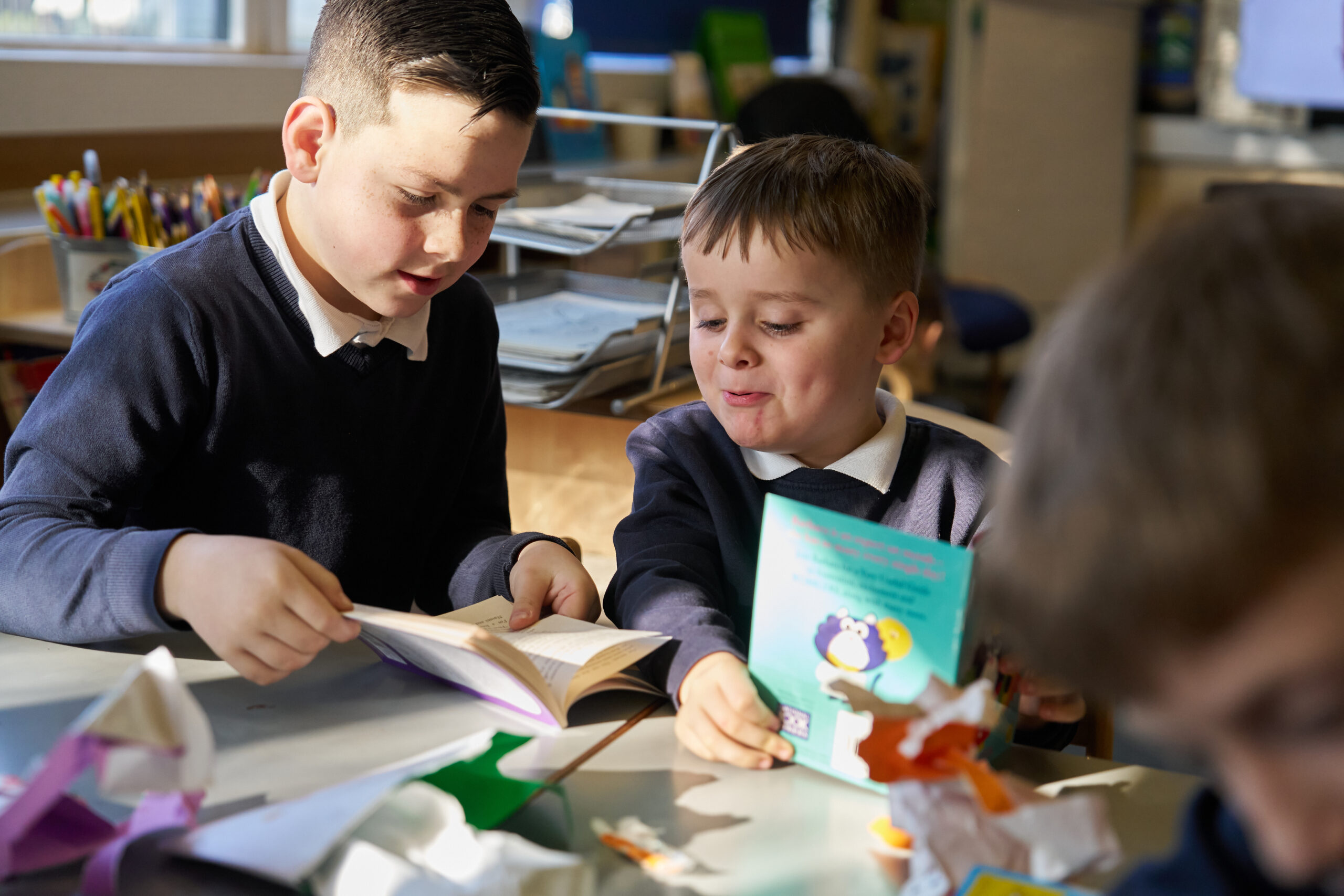Two young boys sit at a table in a classroom, engaged in reading books. One boy holds a book open, while the other smiles and holds a colorful book. The background shows various school supplies and a bright classroom setting.