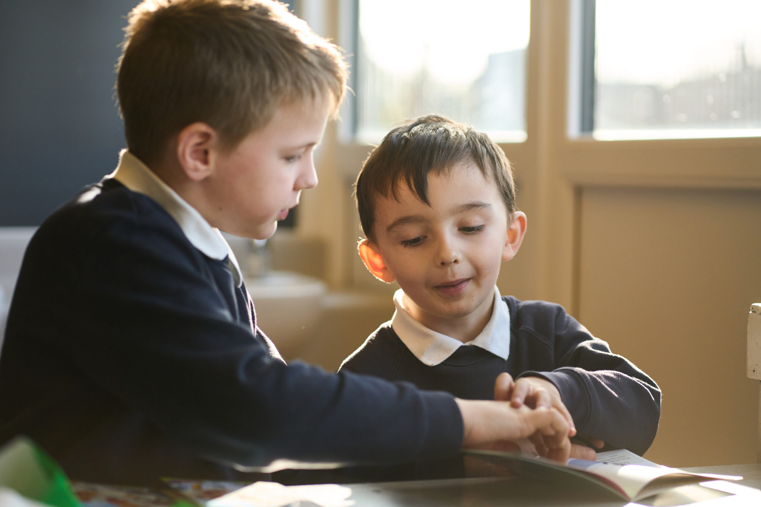 Two young boys are engaged in a focused activity at a table, smiling and interacting as they look at a book together. Natural light streams in from a nearby window, illuminating their expressions.