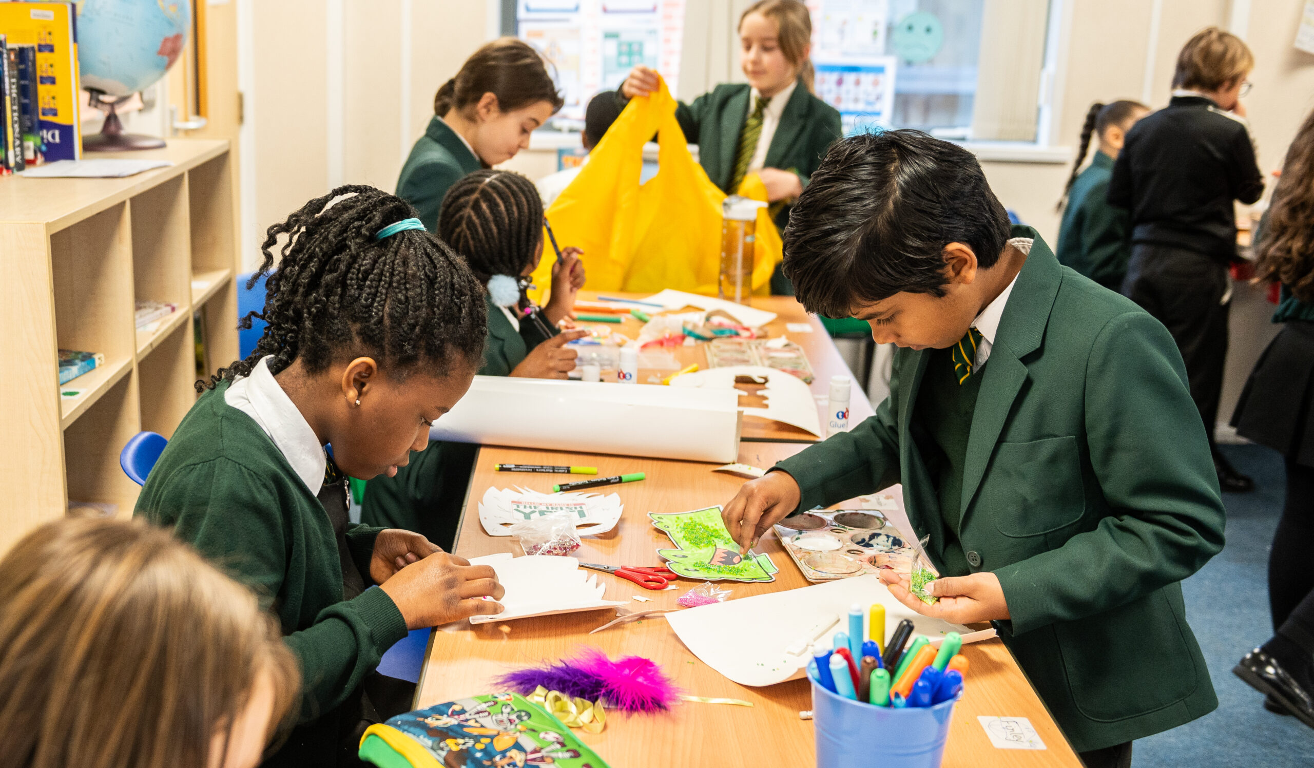 A group of children in school uniforms are engaged in arts and crafts at a table, using scissors, markers, and various colorful materials. Some students are focused on their individual projects, while others are sharing supplies or interacting with their peers.