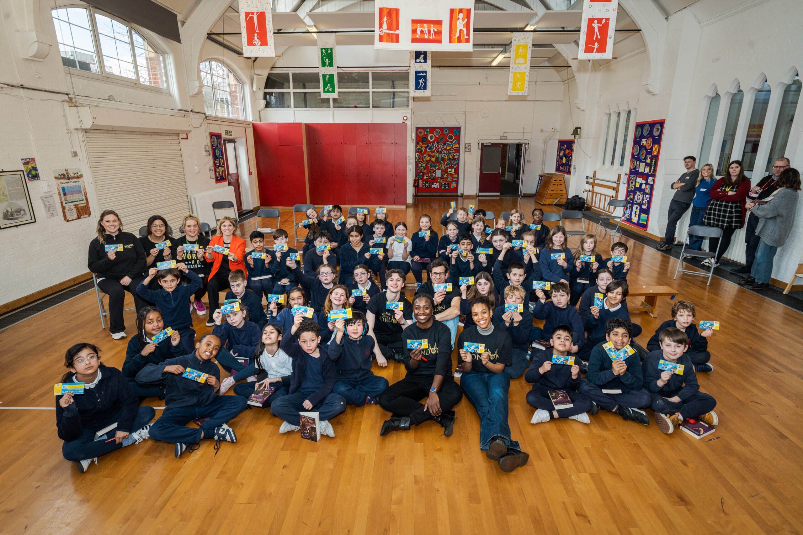 A large group of children in navy sweatshirts smiles while holding up books in a bright, open school gymnasium. Several adults are present in the background, and colorful banners hang from the ceiling, creating a festive atmosphere.