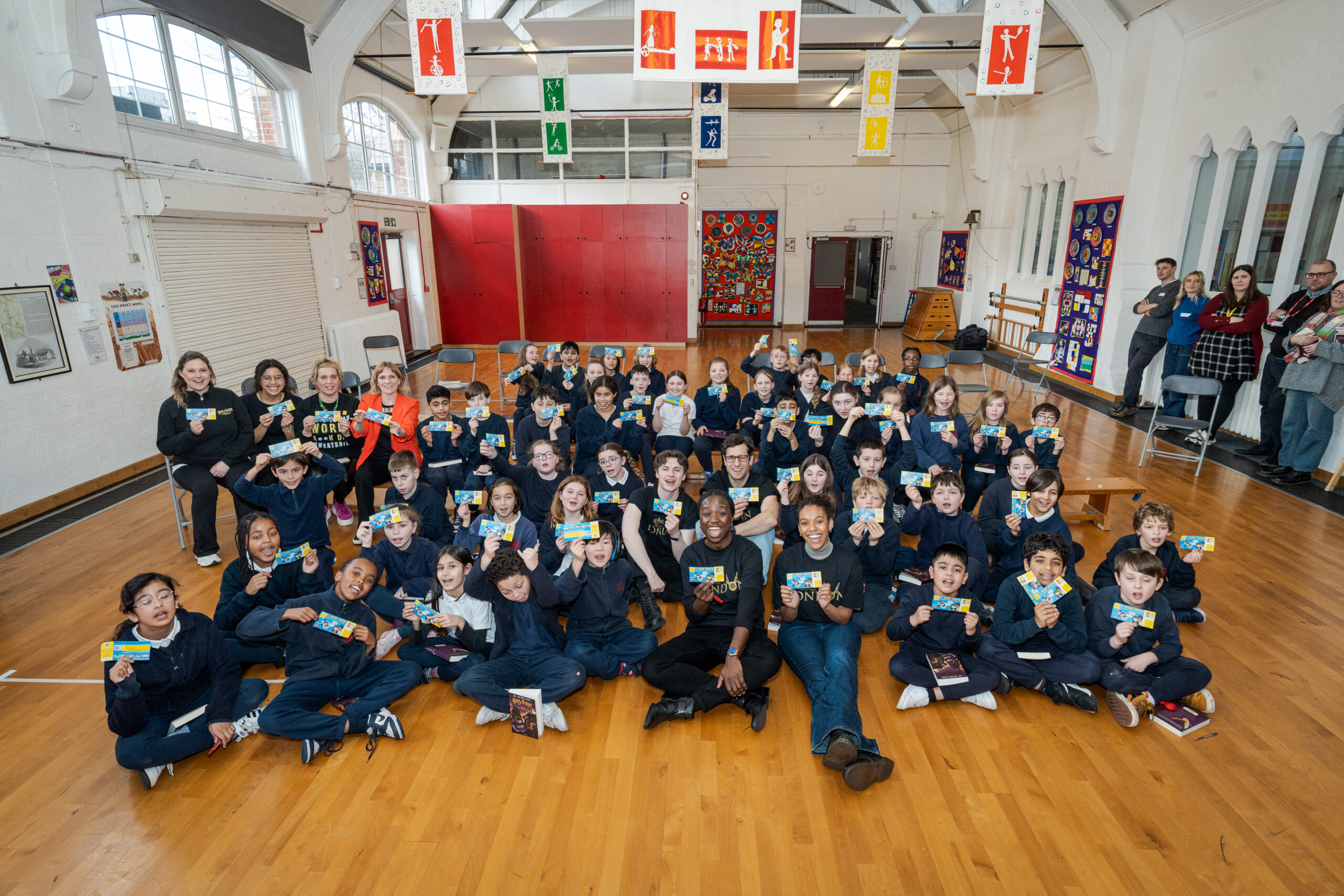 A large group of children, approximately 50, sit on a wooden floor, smiling and holding colorful cards. Two adults are seated among the children, while a few adults stand in the background, all in a bright, spacious room with banners and colorful decorations.
