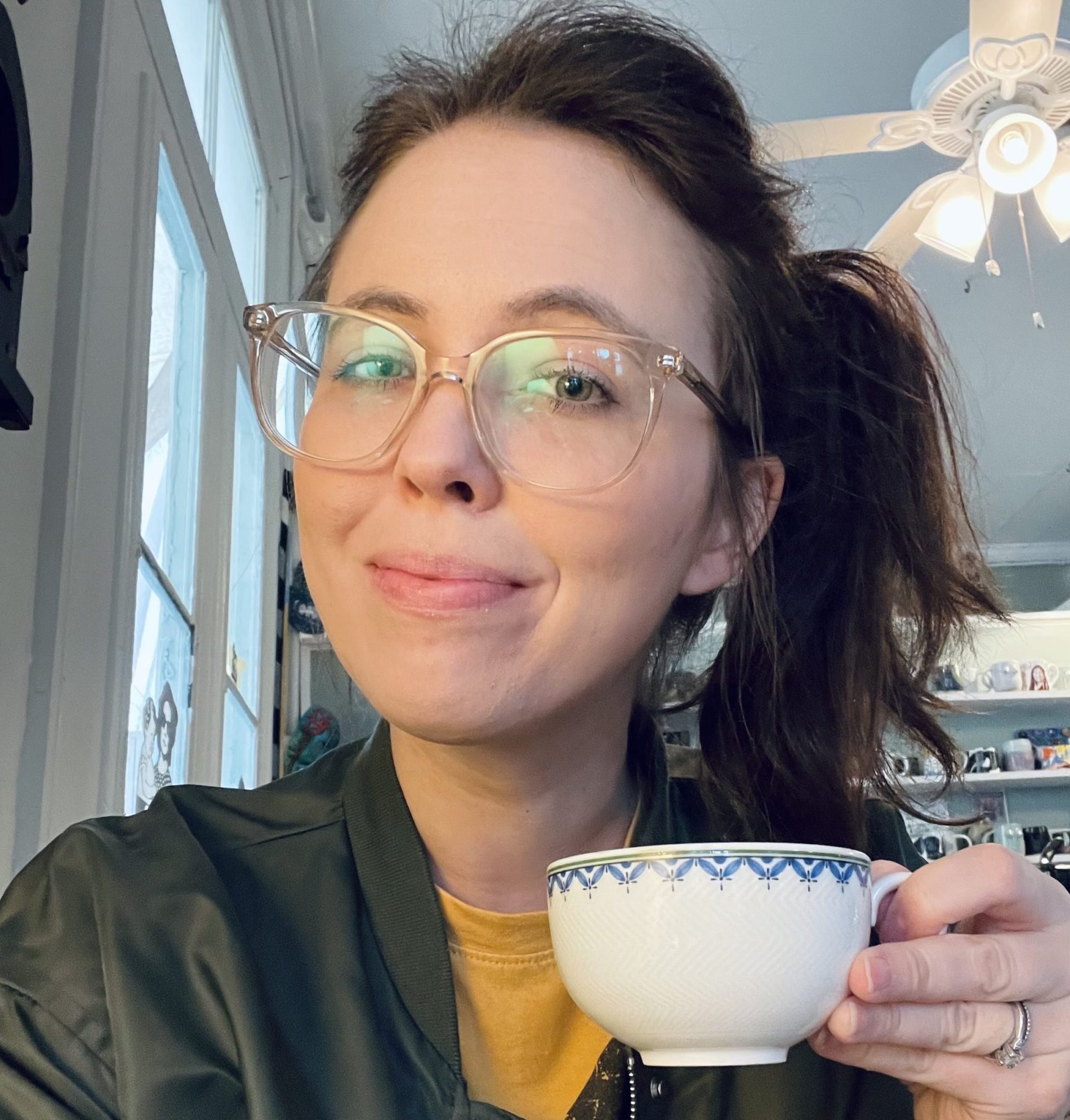 A person with glasses is smiling while holding a decorative cup. They are wearing a dark green bomber jacket and a yellow shirt, sitting in a bright room with shelves visible in the background.