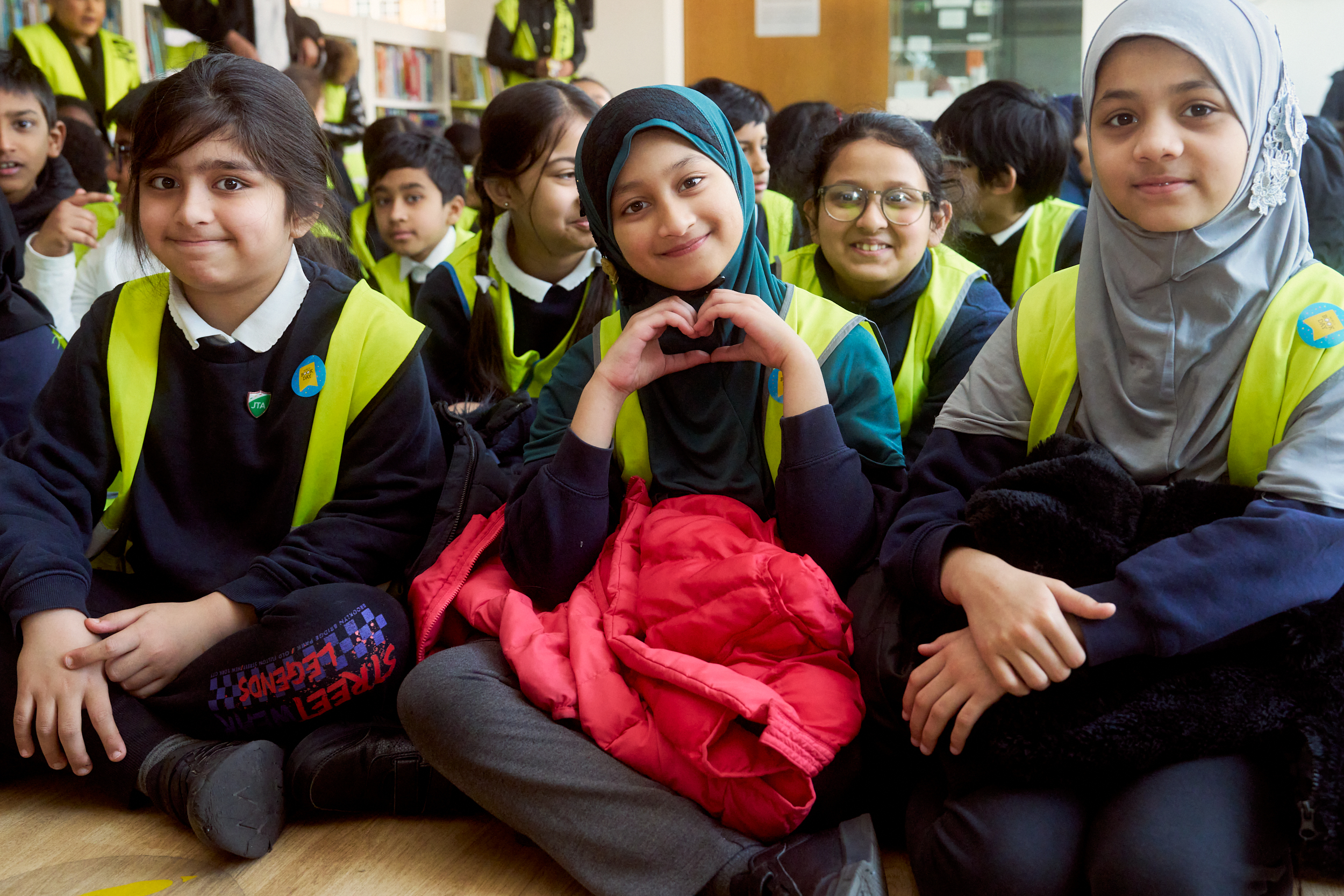A group of smiling children, wearing bright safety vests, sit together on the floor. One girl makes a heart shape with her hands, while others share cheerful expressions, showcasing a sense of friendship and enthusiasm in a classroom setting.