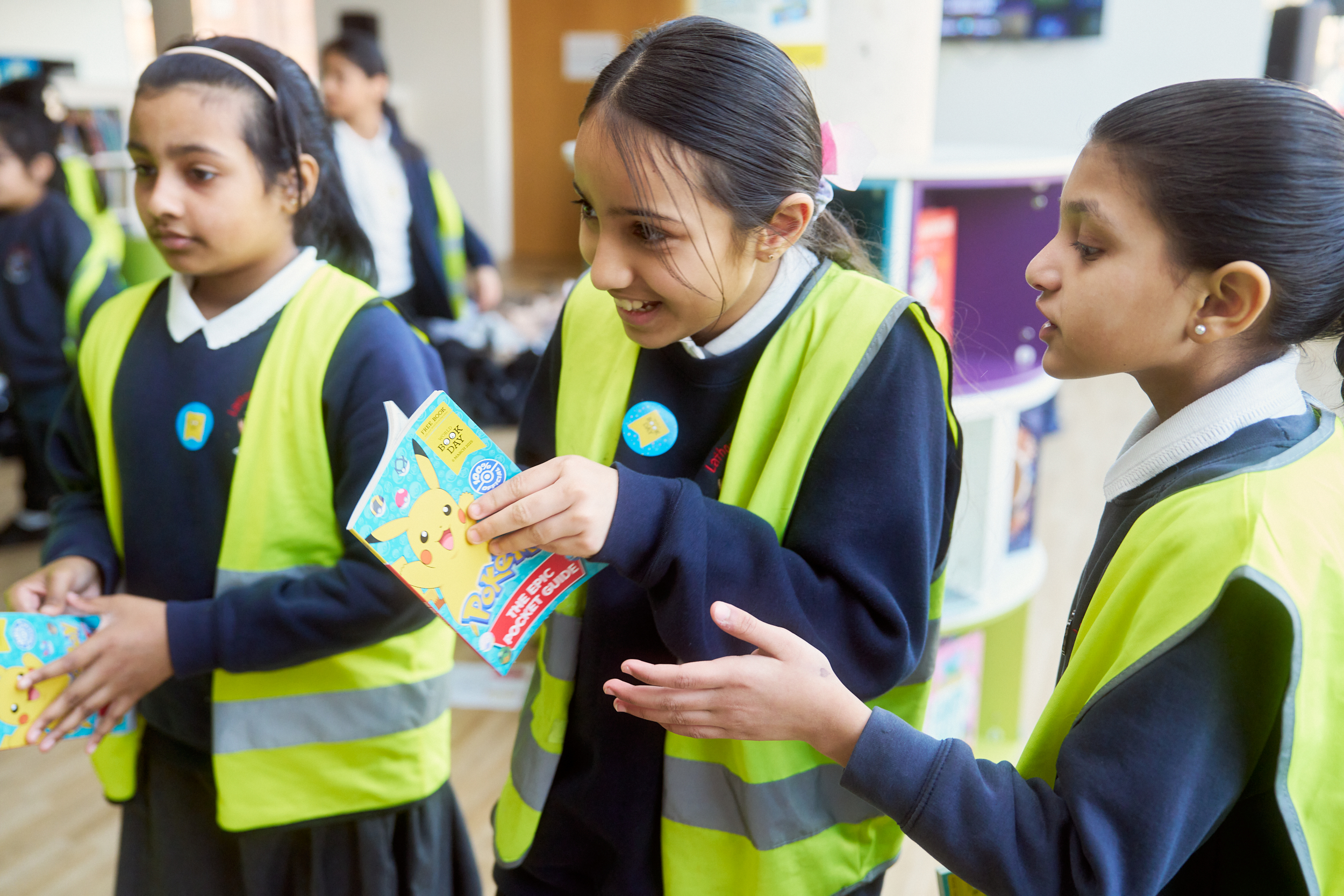 Three young girls wearing bright yellow vests engage in conversation, holding colorful activity books. They appear excited and animated in a classroom setting, with other students in the background. The image captures their enthusiasm for learning and collaboration.