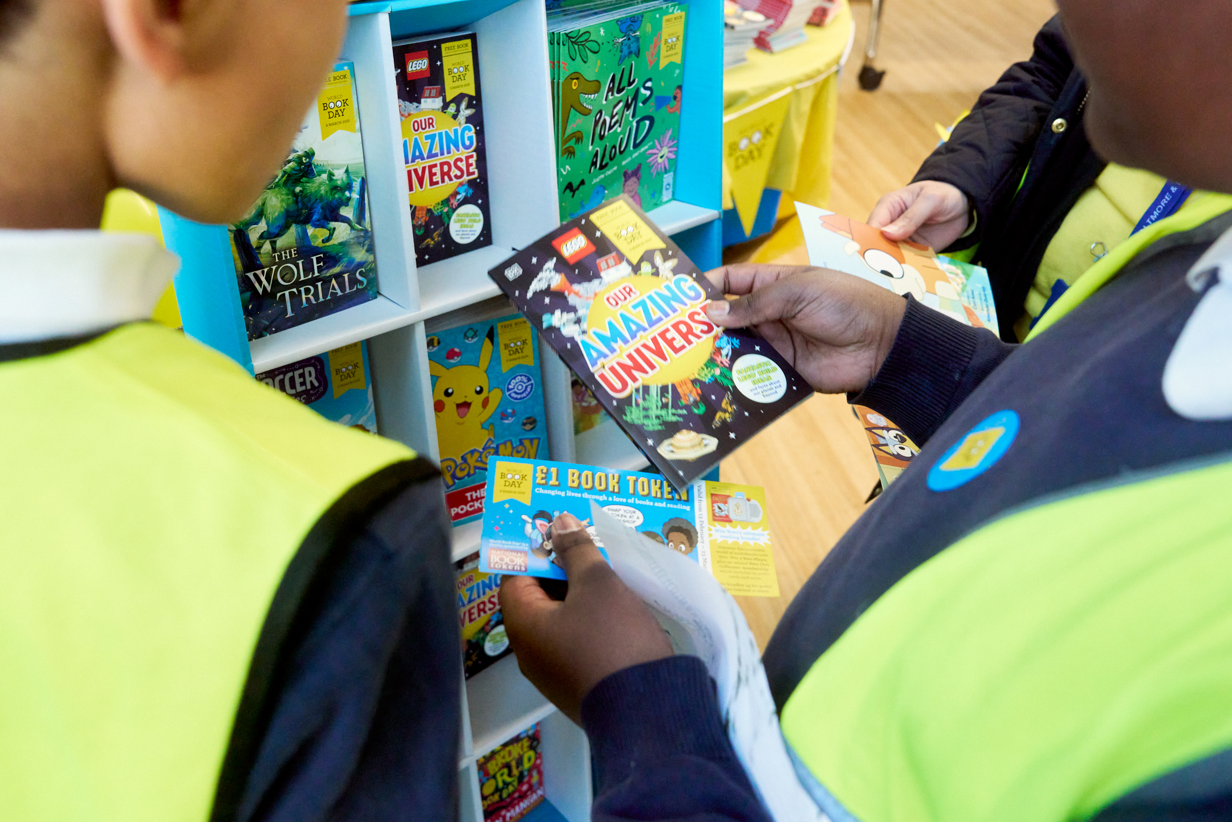 Two children are browsing a colorful bookshelf filled with children's books. One child holds a book titled "Our Amazing Universe" while the other examines nearby titles. Both are wearing bright vests, indicating participation in an educational activity or event.