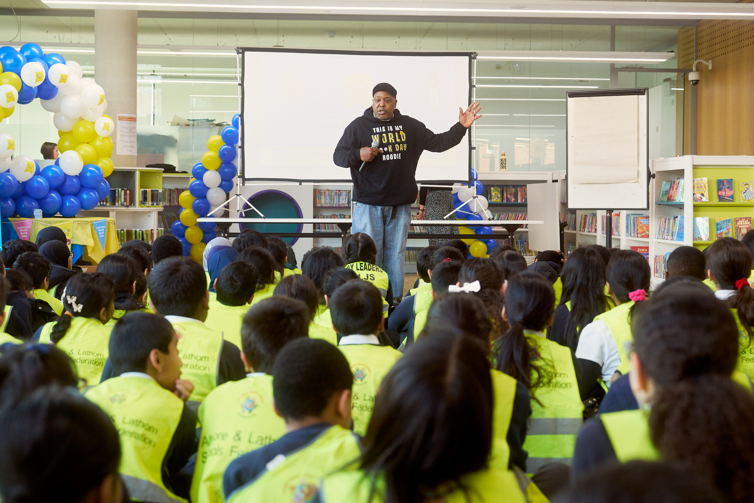 A speaker addresses a group of children wearing bright yellow vests in a library setting, emphasizing engagement and learning. The audience is attentively listening, with a presentation screen visible in the background. Balloons and decorations add a festive atmosphere to the event.