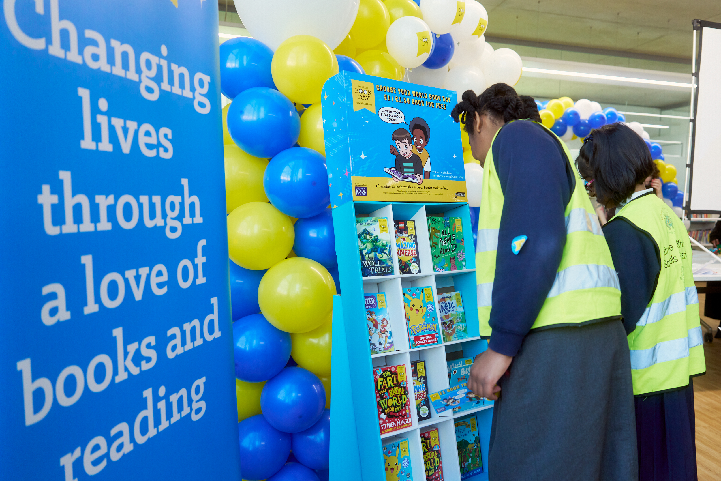 A vibrant library event featuring a colorful display of children's books. Two young individuals in reflective vests browse the selection of books, with a backdrop showcasing a message about the impact of reading. Bright balloons add to the festive atmosphere.