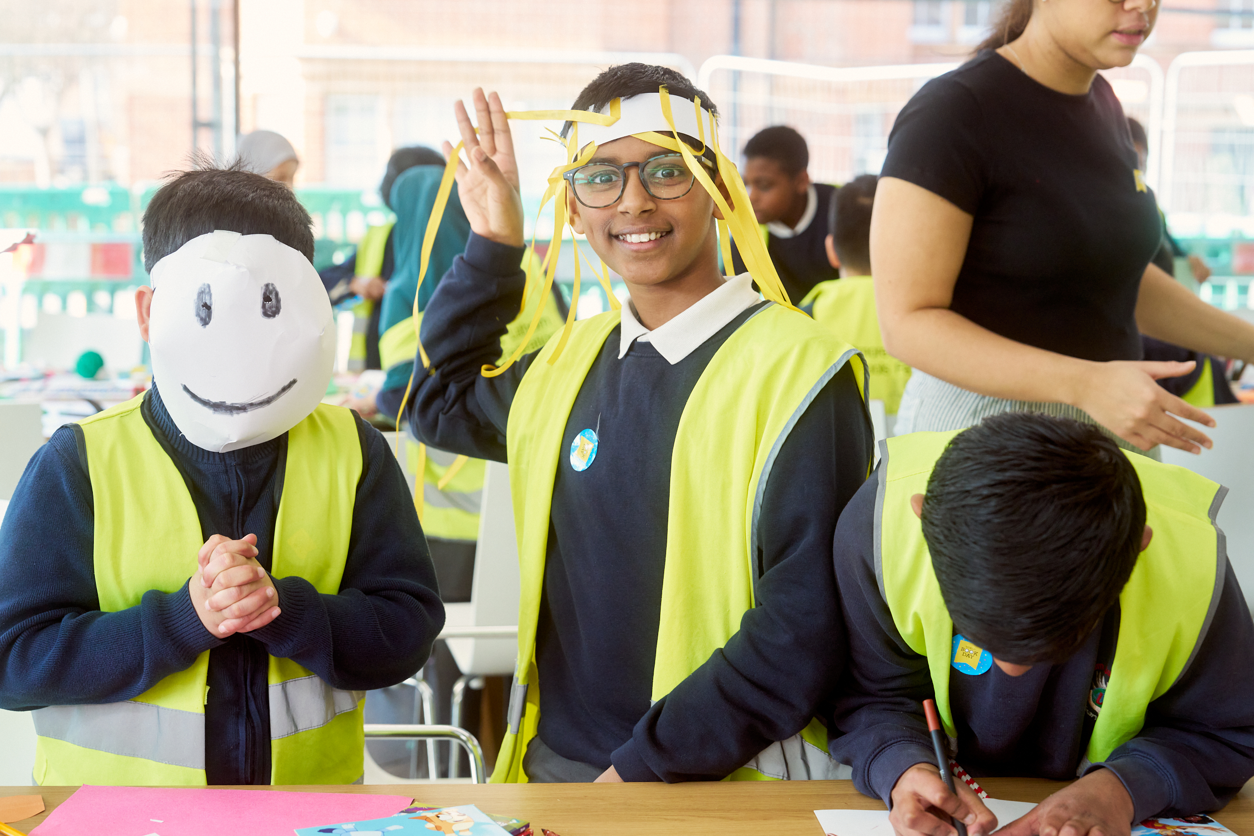 Two children in bright yellow vests engage in a craft activity. One child, wearing a smiley face mask, clasps their hands together, while the other, with glasses and colorful ribbon in their hair, smiles and waves. A teacher assists nearby.