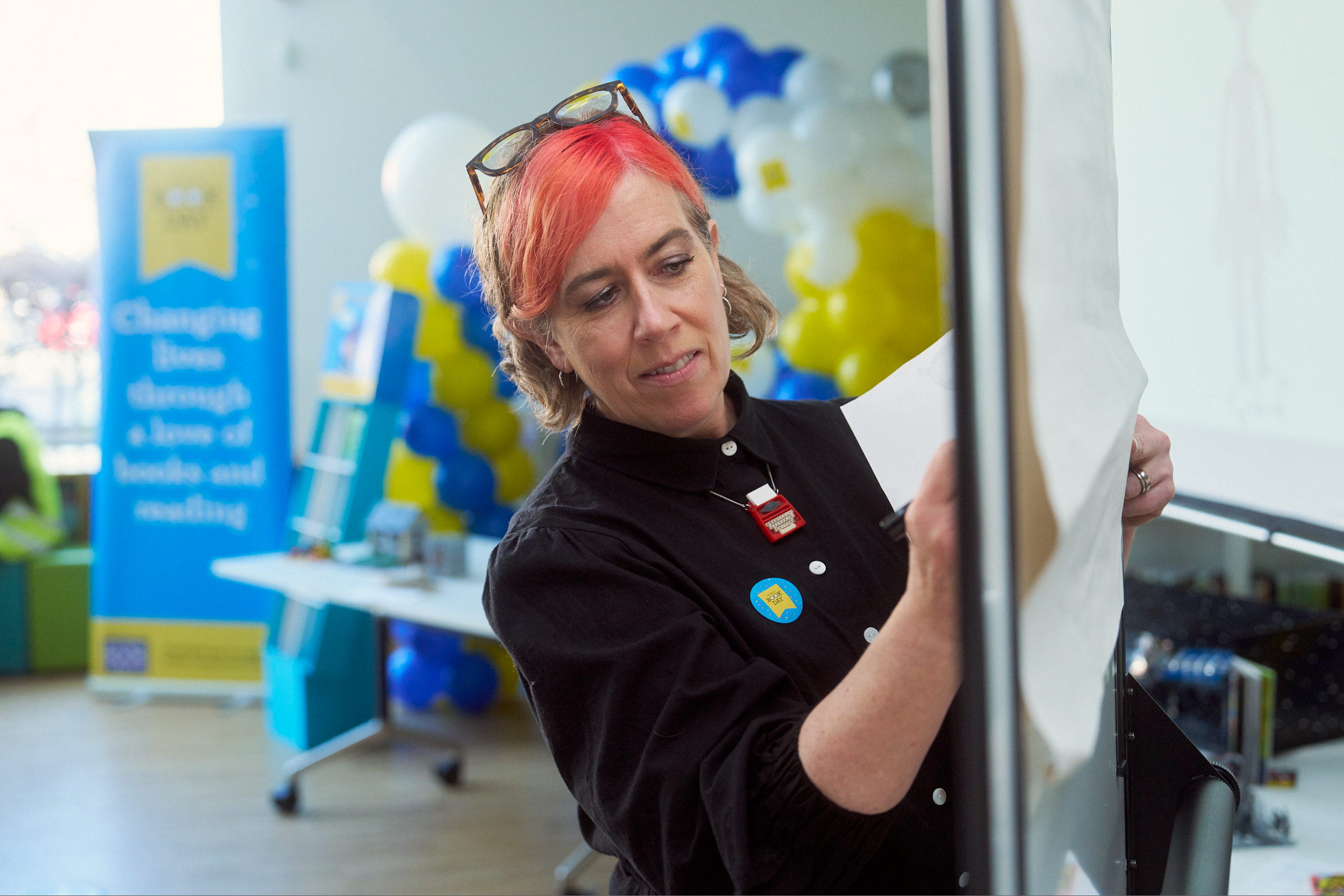 A woman with pink hair is standing in a bright room, engaging with a flip chart. She's wearing a black shirt and a badge, focused on her task. In the background, colorful balloons and a banner promoting literacy are visible.