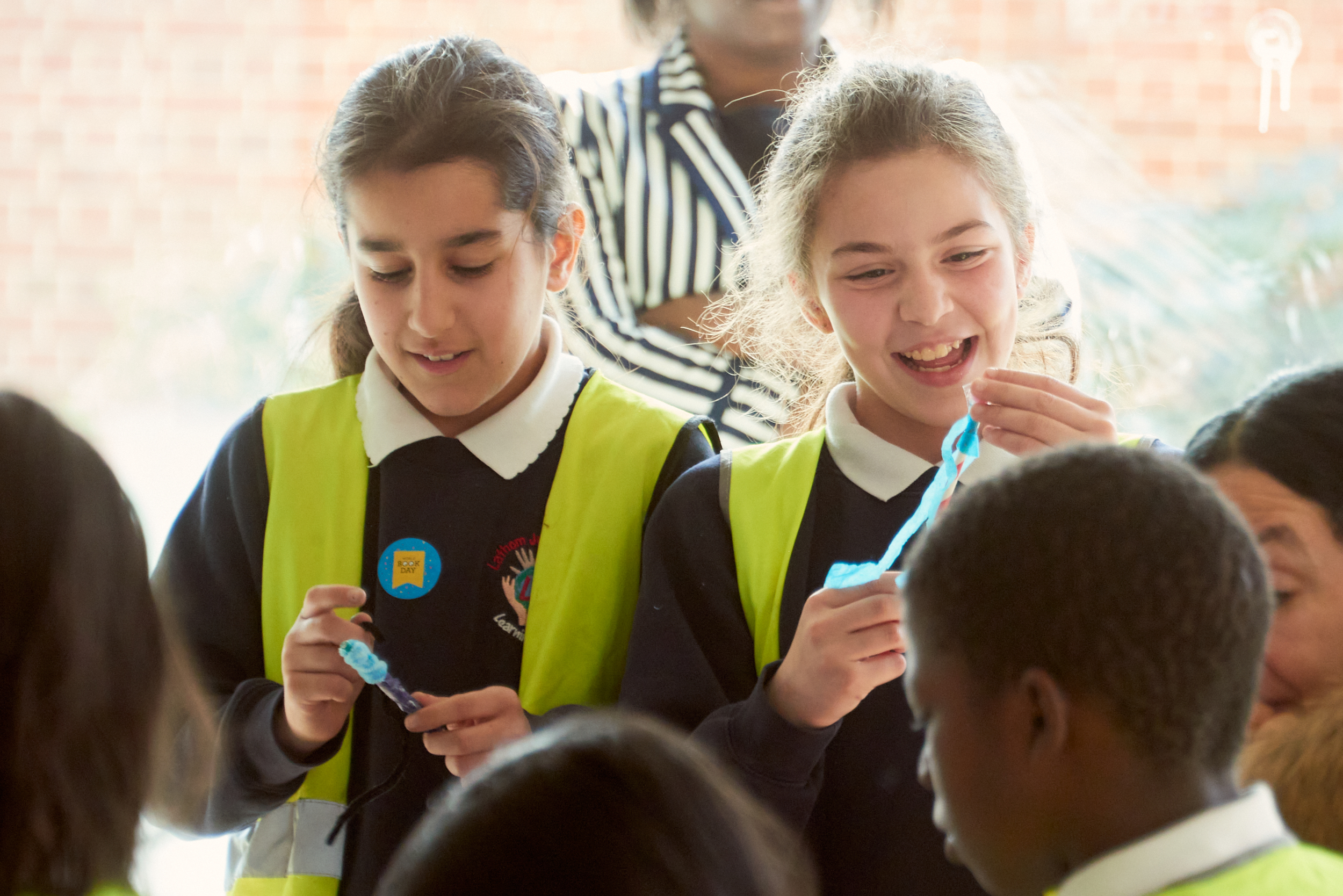 Two smiling girls in safety vests hold colorful materials, engaged in a fun activity. In the background, other children and adults are gathered, creating a lively atmosphere.