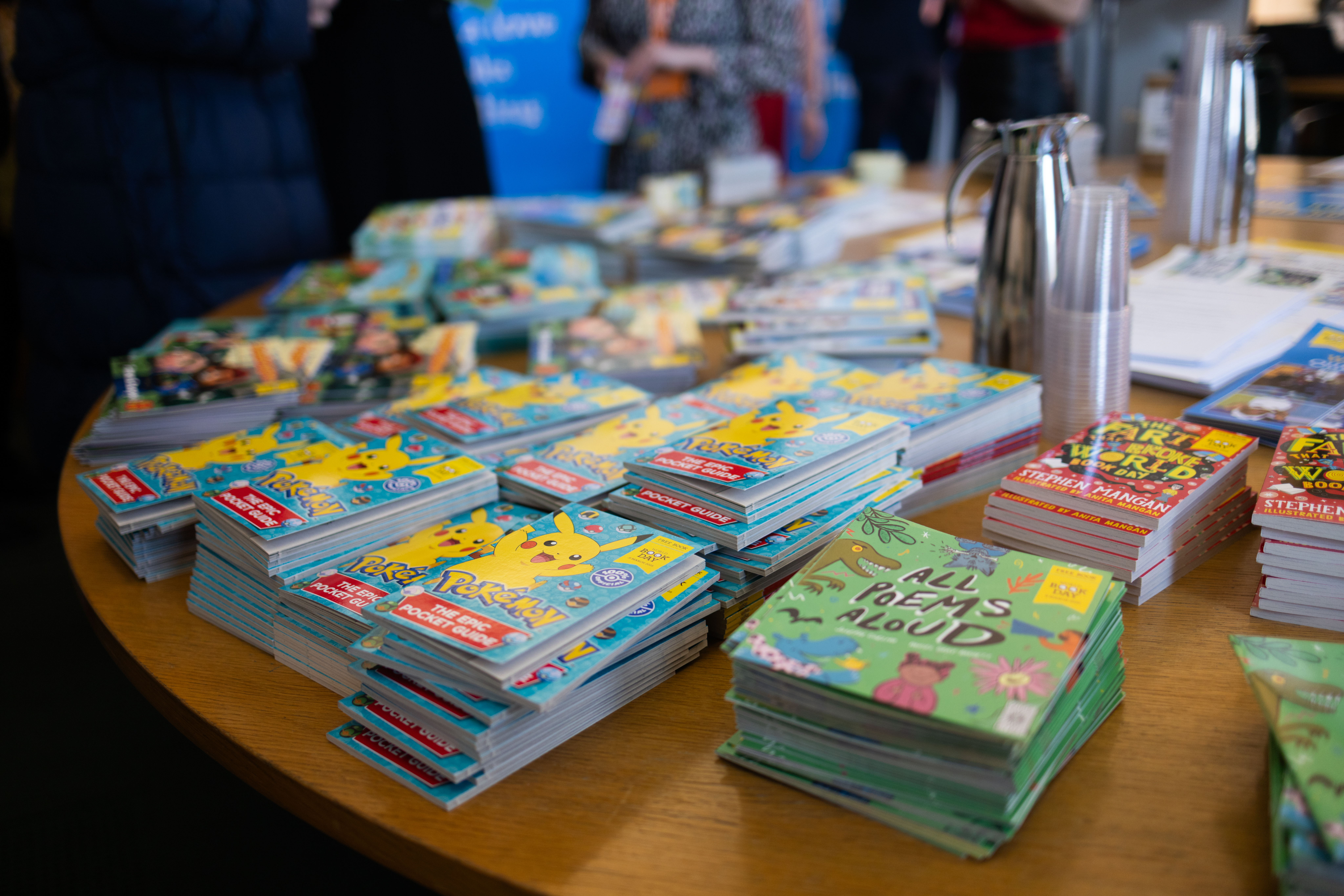 A table covered with colorful children's books and pamphlets, including titles featuring Pokémon and poetry. Nearby, a coffee pot and cups suggest a gathering or event. People can be seen in the background, indicating a communal atmosphere focused on reading.