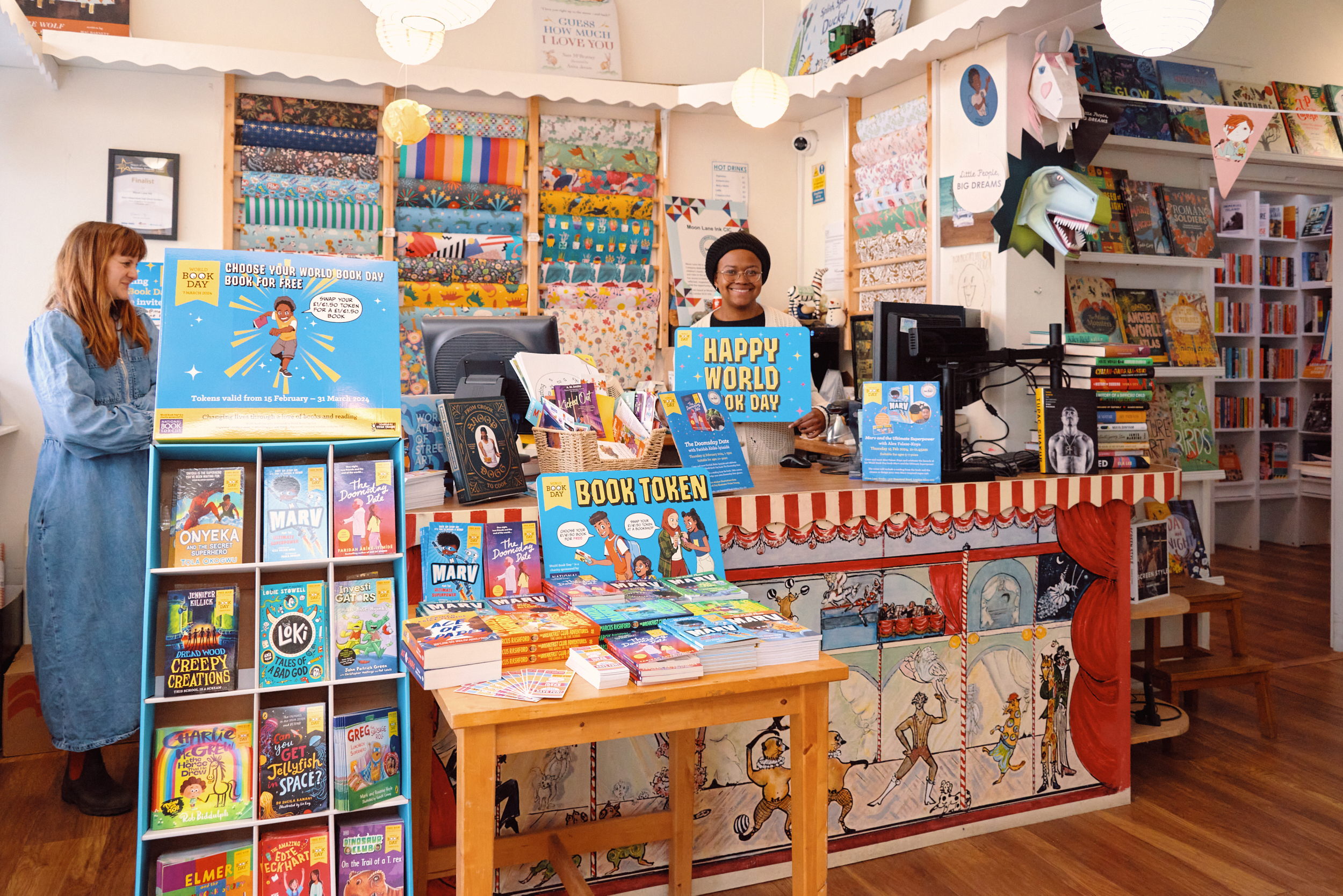 A vibrant bookstore interior features a smiling employee at the counter, celebrating World Book Day. Shelves are filled with colorful books, and a display promotes book tokens. The space is decorated with playful artwork and bright colors, creating an inviting atmosphere for readers.