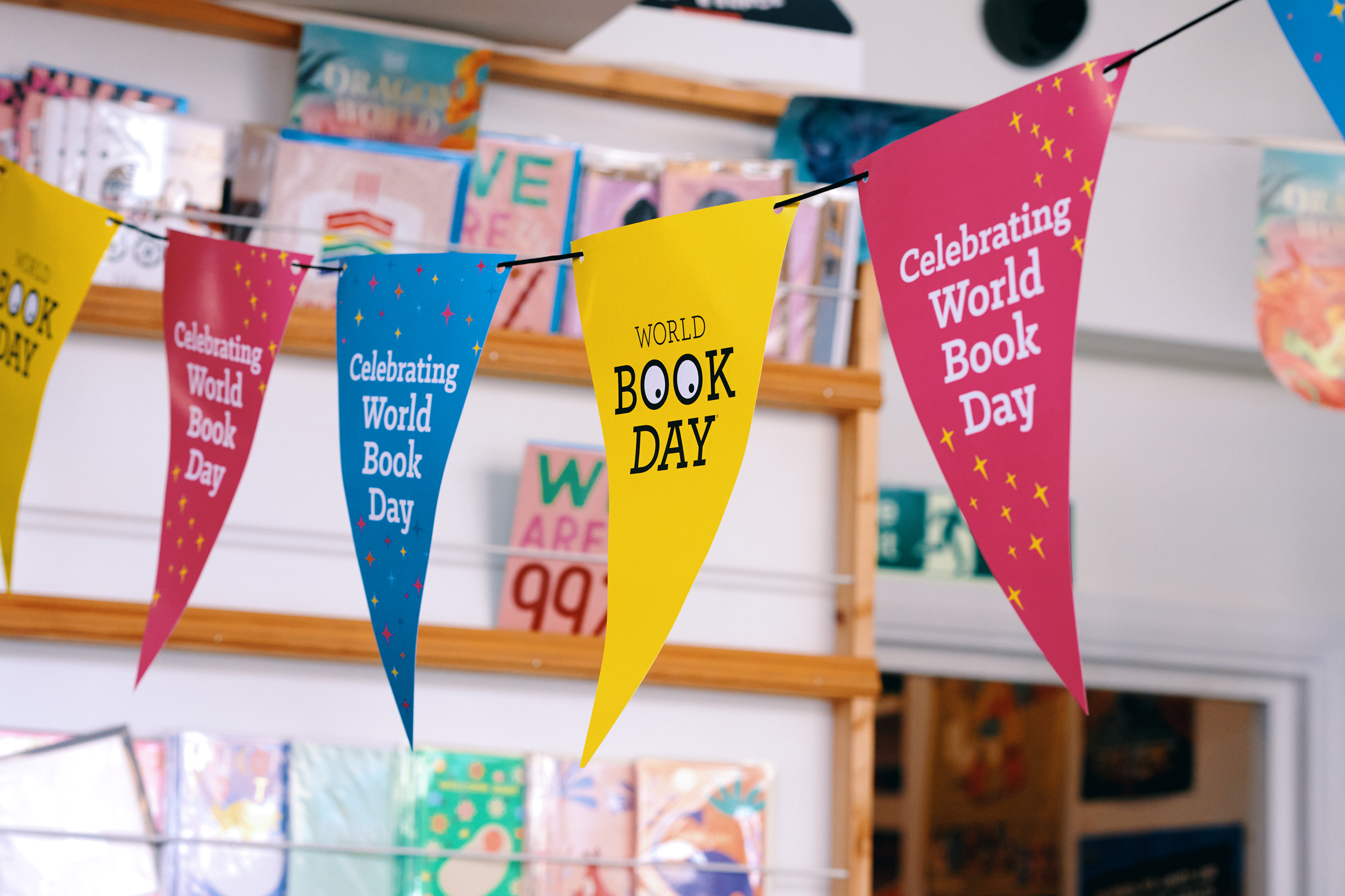 Colorful pennants celebrate "World Book Day," featuring messages like "Celebrating World Book Day." The background shows various books and decorative elements, creating a festive atmosphere in a bookstore setting.