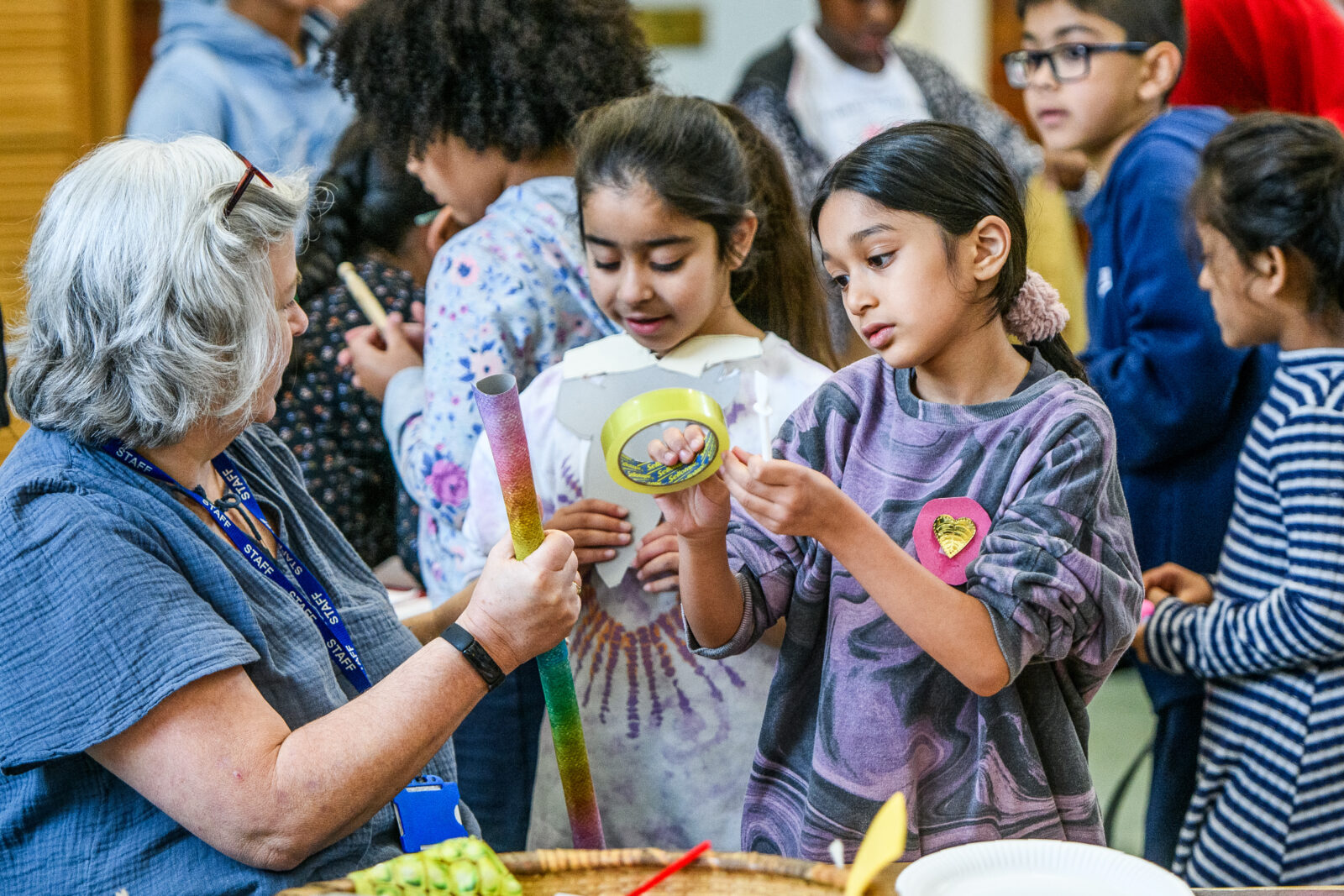 A group of children and an adult engage in a hands-on activity, exploring arts and crafts materials. The adult shows a colorful tube while a girl inspects a decorated bowl, surrounded by other children. The atmosphere is lively and creative, promoting collaboration and learning.