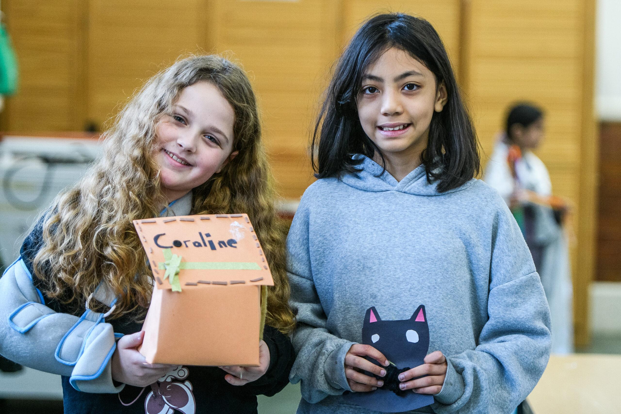 Two girls smile while holding craft projects. One girl has long curly hair and holds an orange box labeled "Coraline," while the other girl wears a gray hoodie and holds a black cat figure. The background features a classroom setting with wooden panels.