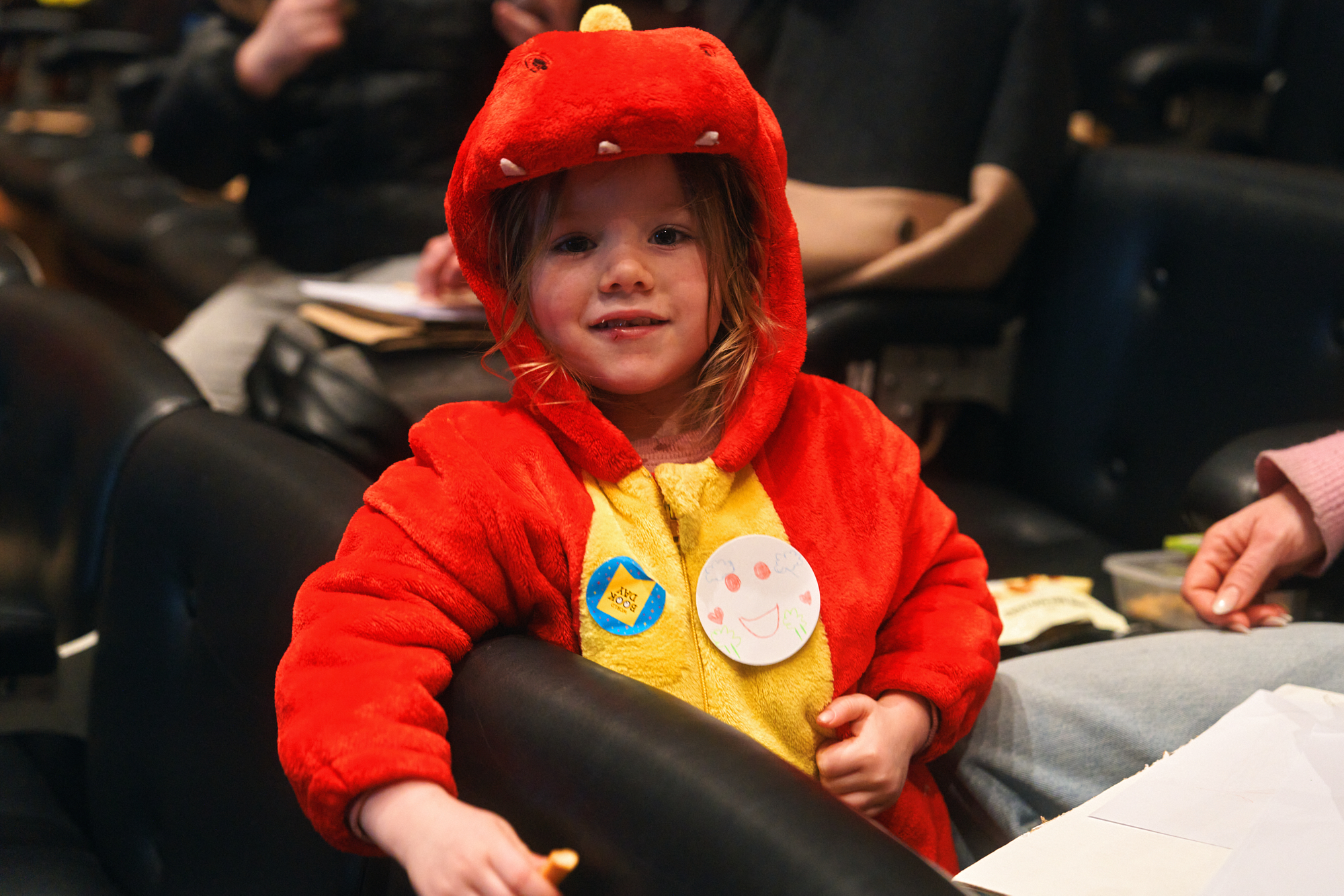 A happy child wearing a bright red dinosaur costume sits in a chair, smiling. They have two stickers on their costume and are surrounded by people in a casual setting.