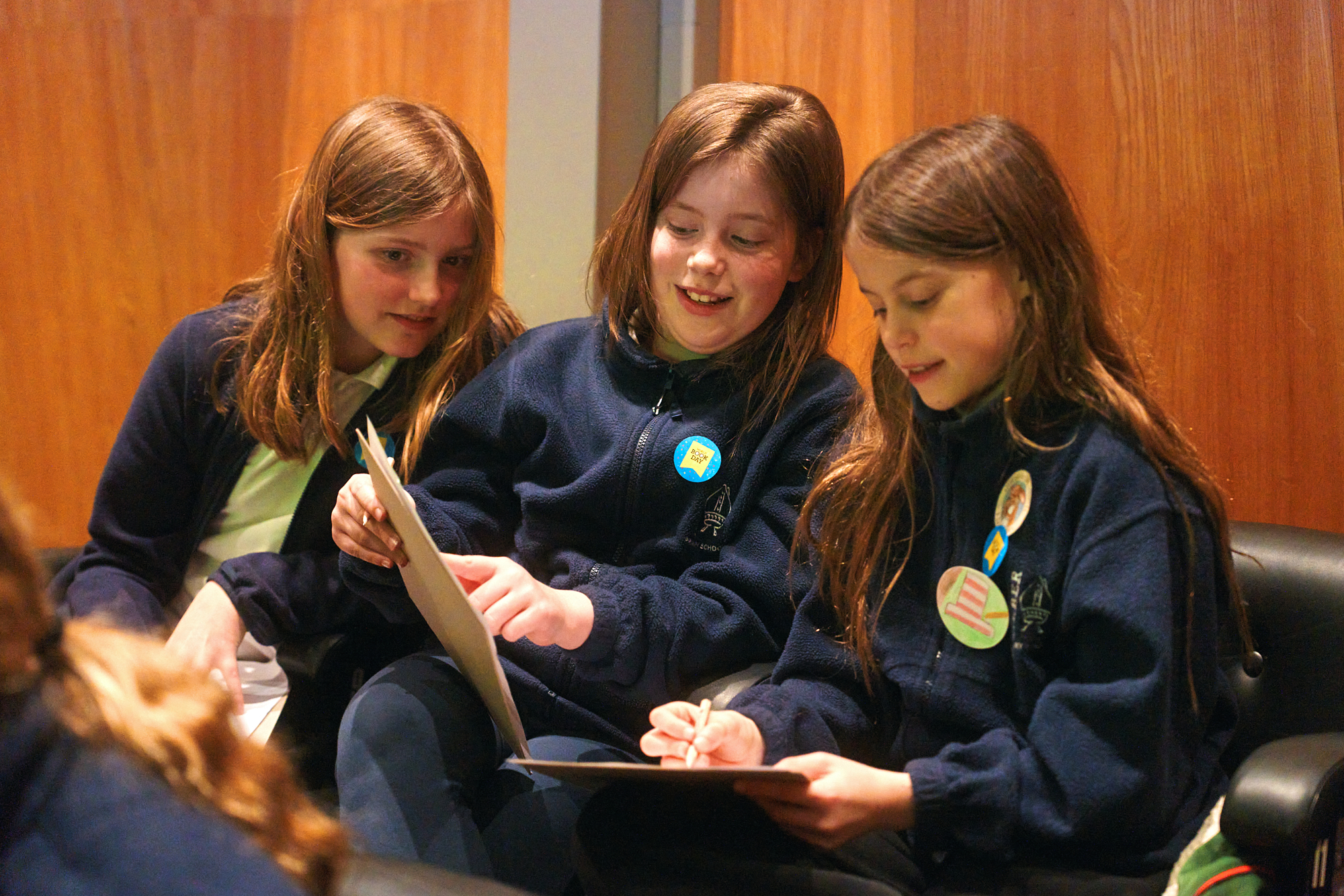 Three girls are sitting together, smiling and engaged in an activity. They are holding papers and writing, wearing matching navy sweatshirts, and have colorful badges on their clothing. The background features wooden panels, creating a warm and inviting atmosphere.