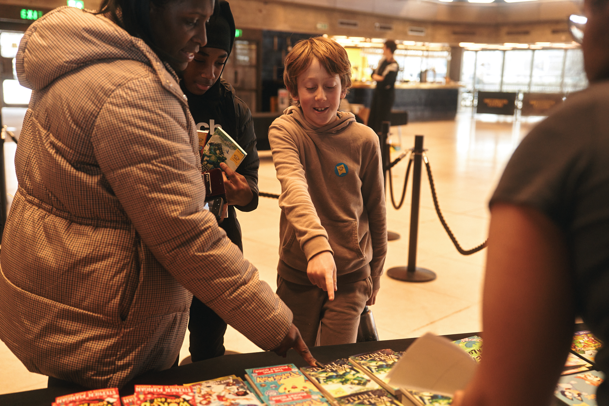 A woman and a boy are engaged in a discussion over a table filled with books. The boy, with short brown hair, points at the books while looking intrigued. Another child stands nearby, holding a book. The scene takes place in a spacious indoor setting.