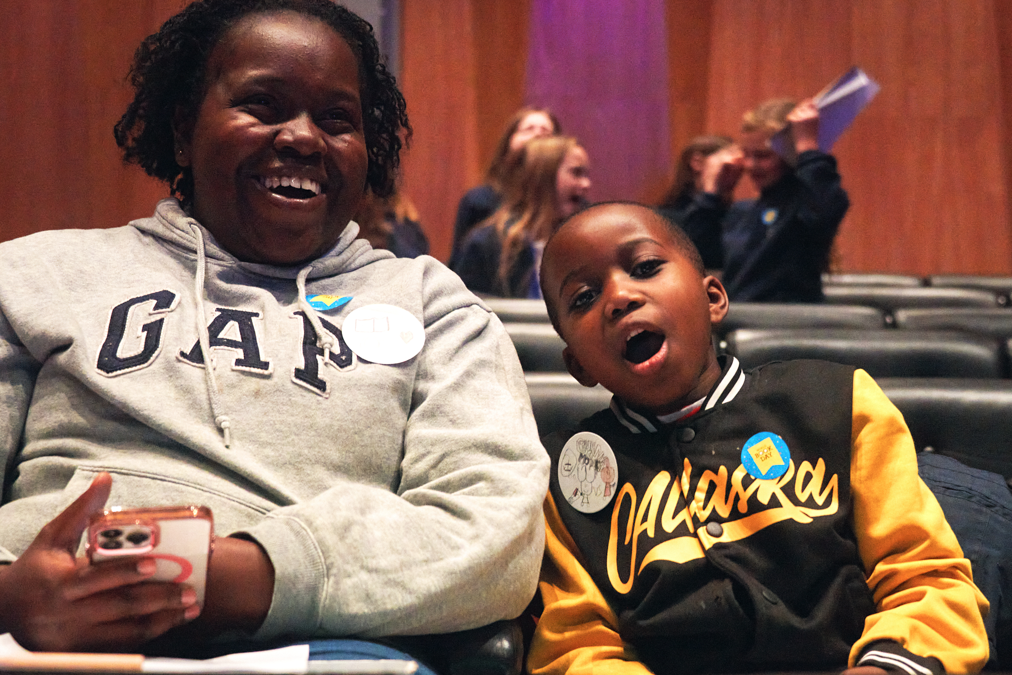 A joyful woman and a young boy sit together in a theater, both smiling widely. The woman wears a gray hoodie, while the boy is dressed in a black and yellow jacket. They appear to be enjoying an event, surrounded by other attendees in the background.