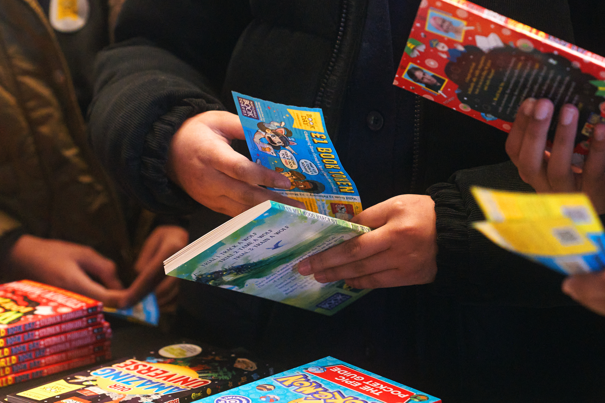 A group of people sorts through various books at a table, with one person holding a colorful book and flyers. The scene captures the excitement of browsing and selecting reading materials.