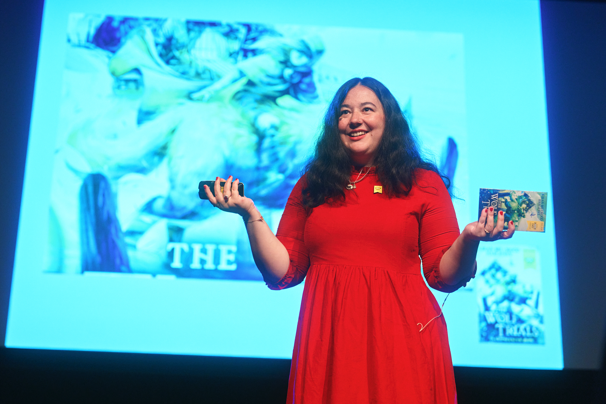 A speaker in a red dress holds up a book while presenting on stage. A colorful illustration is displayed behind her, highlighting key themes from her talk. The atmosphere is engaging, with the audience focused on the presentation.