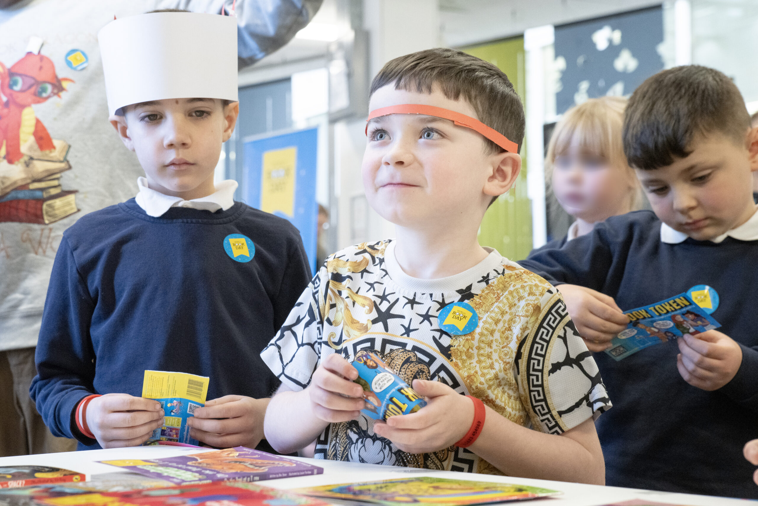 A group of young children in a classroom setting are engaged in a fun activity. One boy, wearing a colorful shirt and a headband, looks excited while holding a small item. Two other children beside him are also focused on their activities, showcasing the vibrant learning environment.