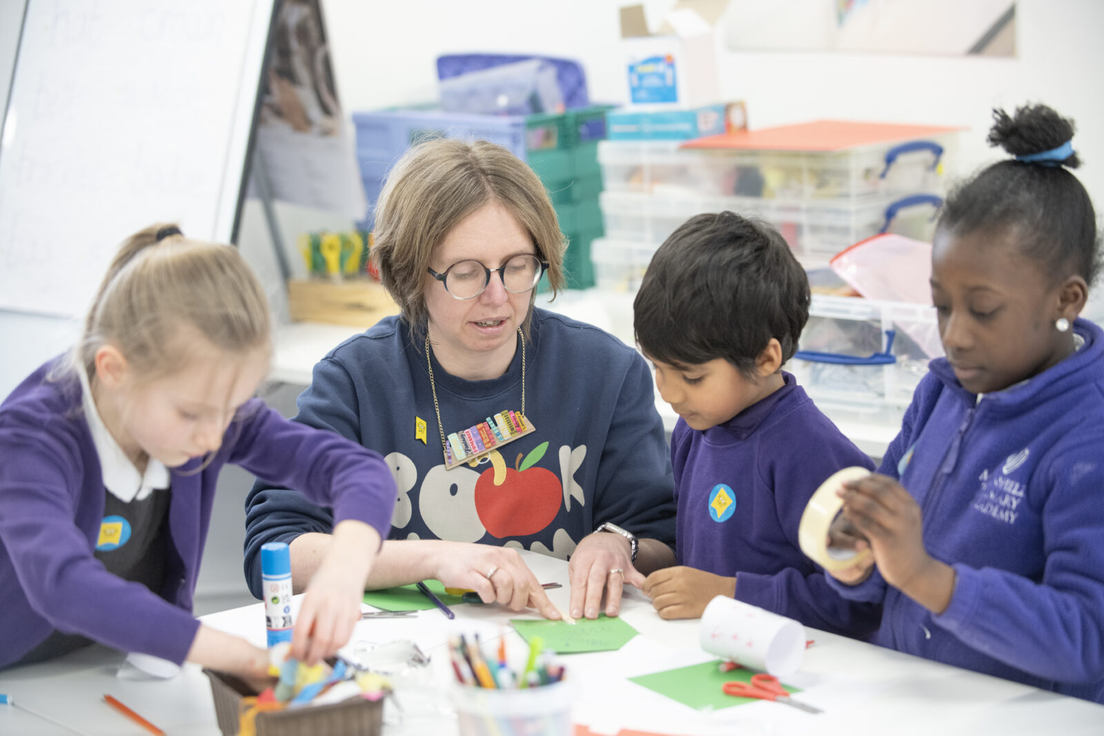 A teacher guides three children in a colorful classroom as they engage in a hands-on craft activity, using paper and adhesive materials to create artistic projects. The atmosphere is creative and collaborative, with various craft supplies visible on the table.
