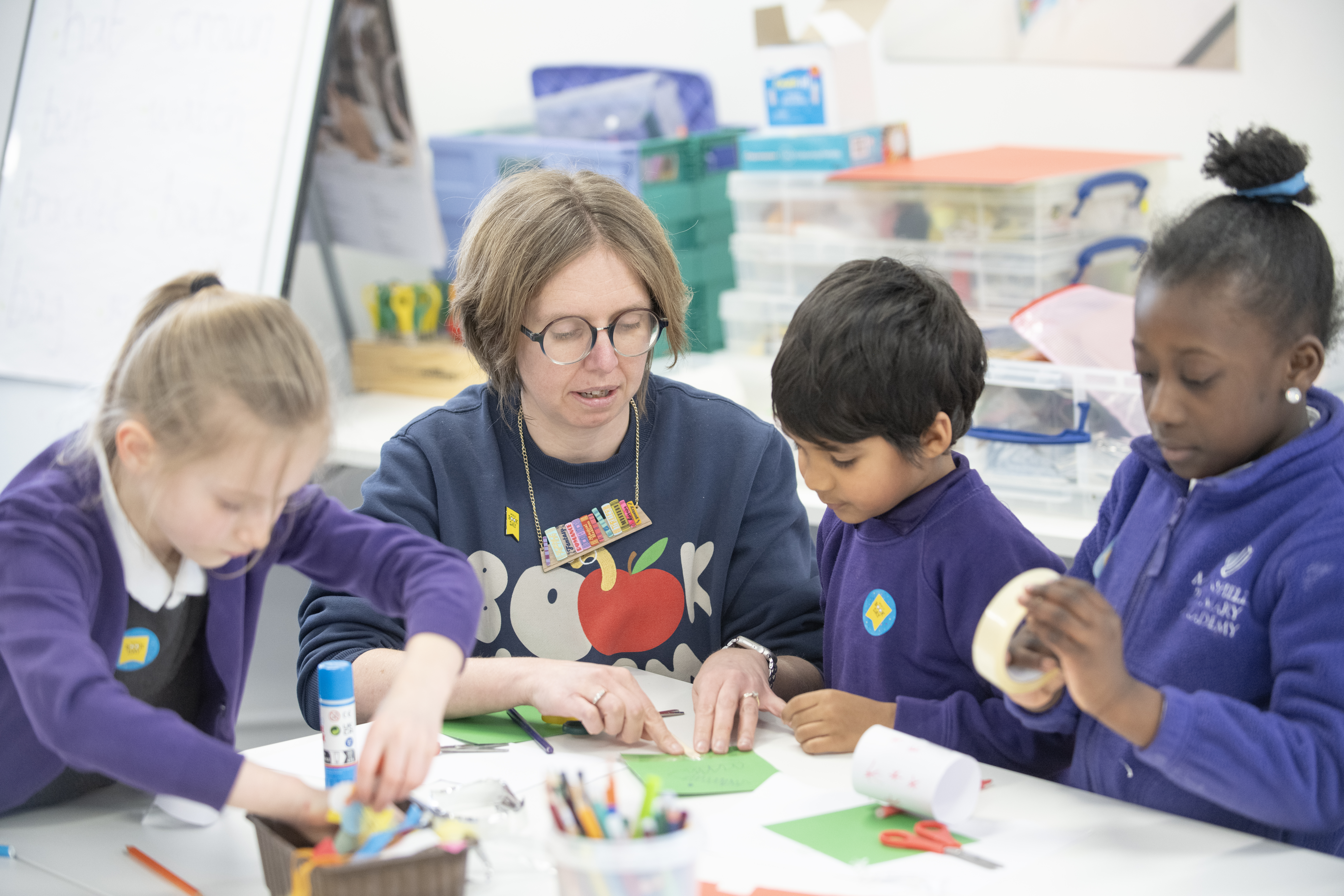 A teacher guides three children in a colorful classroom as they engage in a hands-on craft activity, using paper and adhesive materials to create artistic projects. The atmosphere is creative and collaborative, with various craft supplies visible on the table.