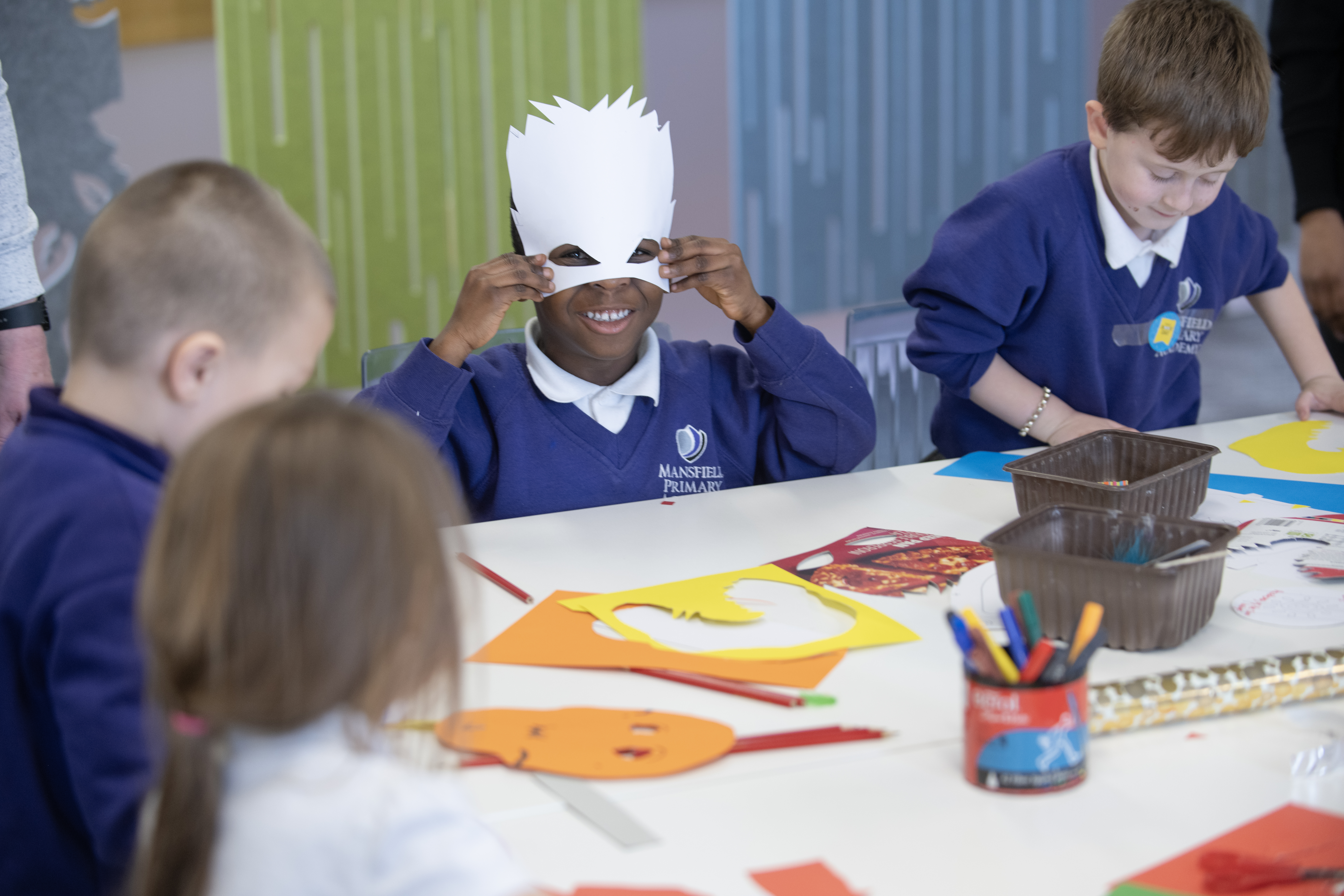 A group of children engages in an art activity, with one child wearing a paper mask. The table is covered with colorful paper cutouts and art supplies, creating a lively, creative atmosphere.