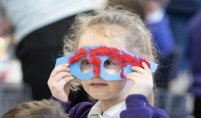 A young child wearing a colorful, handmade mask with red and blue decorations holds it up to their face. They are in a classroom setting, surrounded by other children engaged in activities. The child is dressed in a purple cardigan and a white shirt.