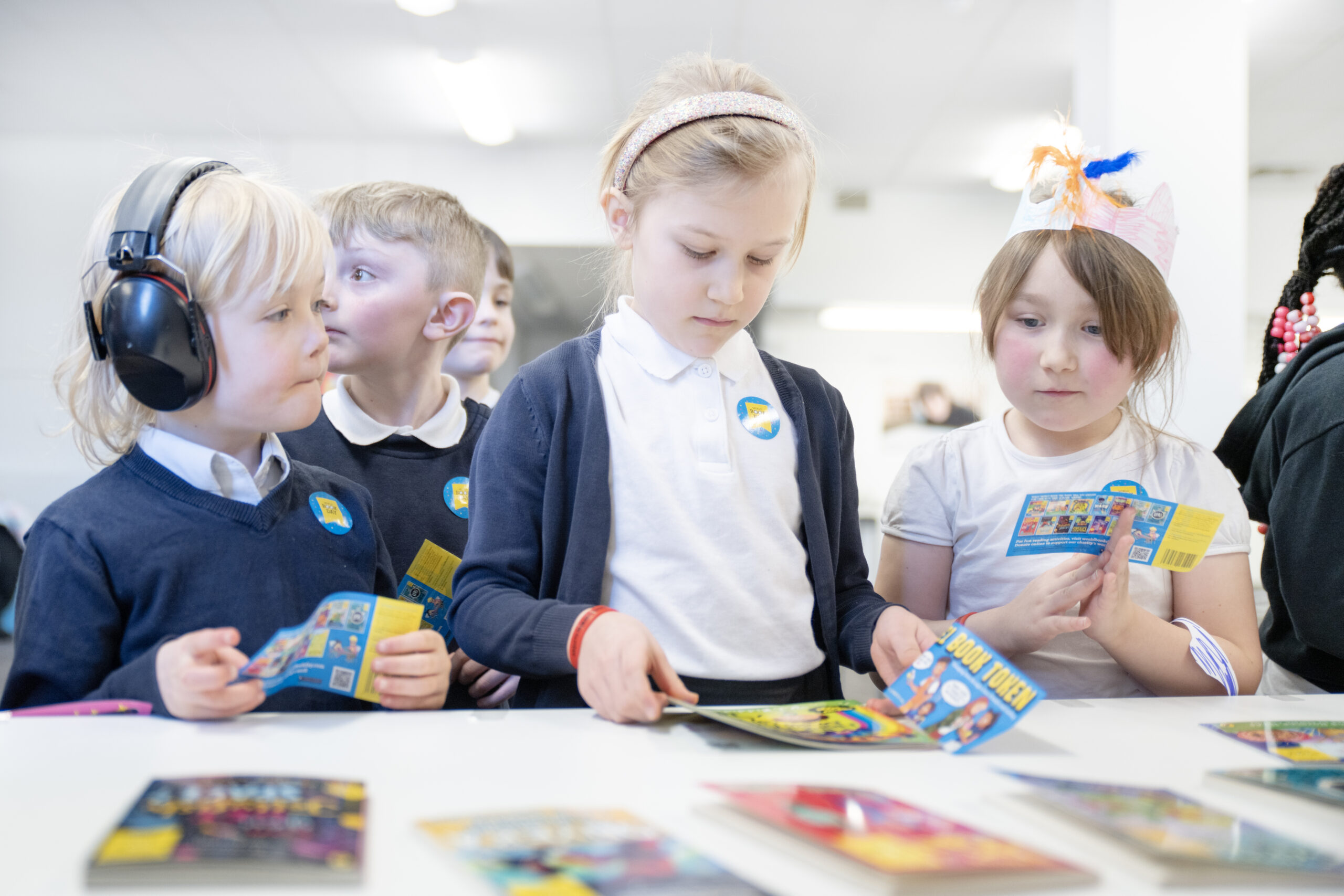 A group of young children are engaged in reading colorful brochures at a table. One child wears headphones, while another examines a brochure. They are in a bright, minimalistic setting, focused on their activities.