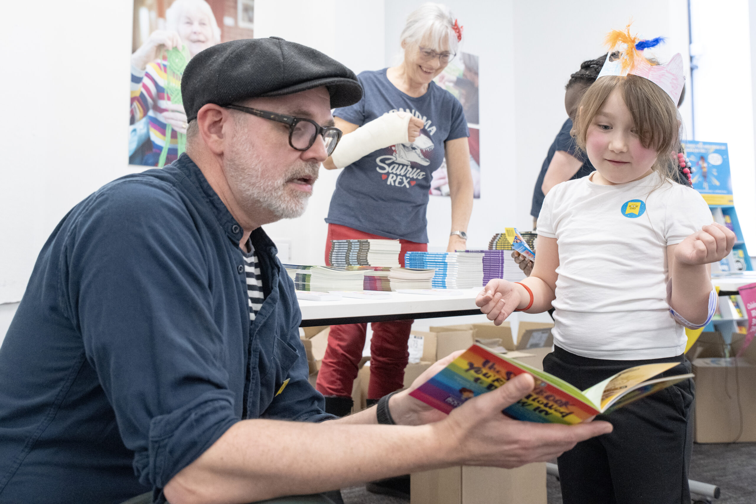 A man in a cap reads a colorful book to a young girl, who looks intrigued. In the background, a woman with a bandaged arm is engaged with other participants. Books are stacked on a table, creating a lively reading environment.